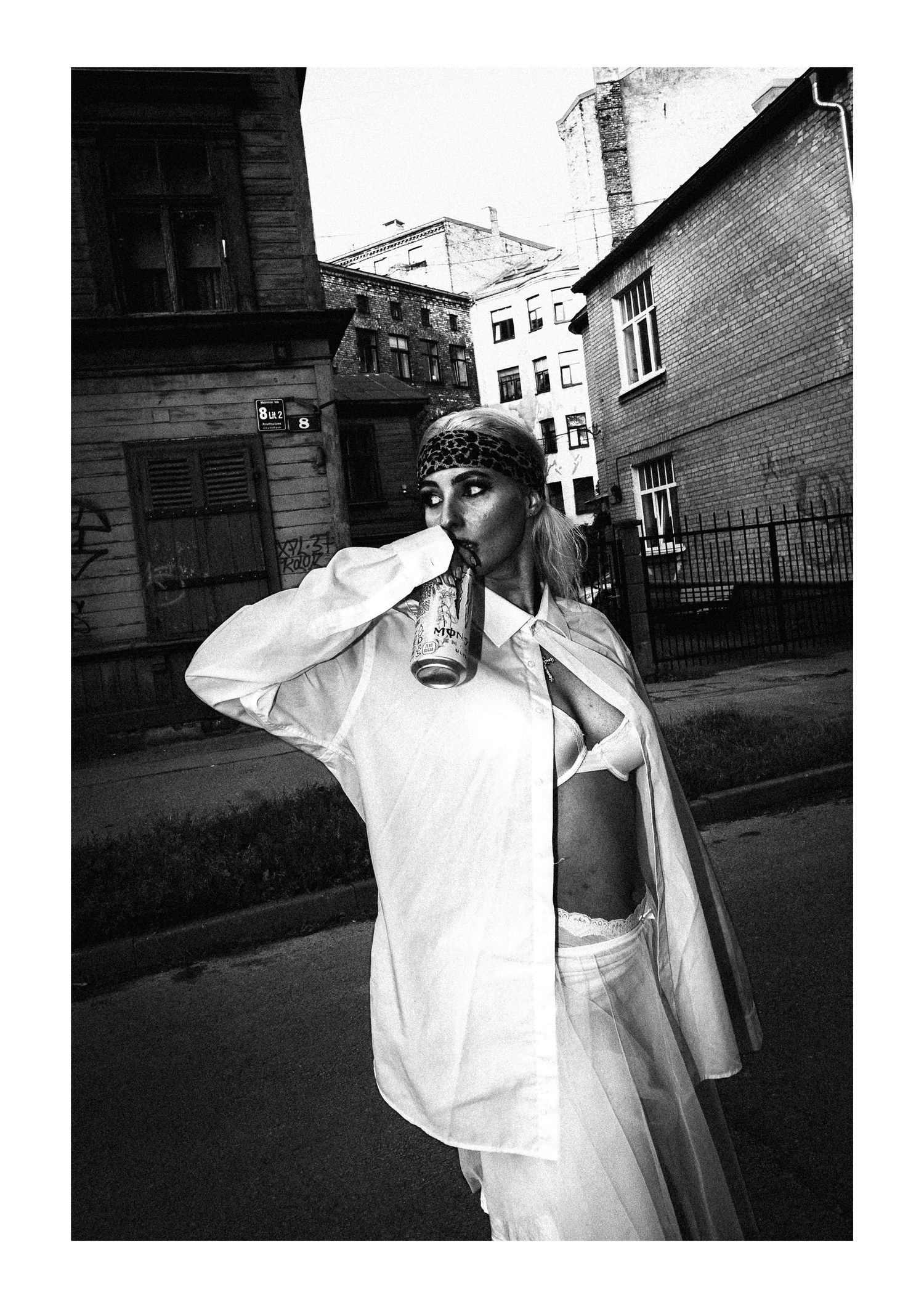 A figure in a white oversized outfit and sunglasses drinks from a bottle on an urban street in black and white.