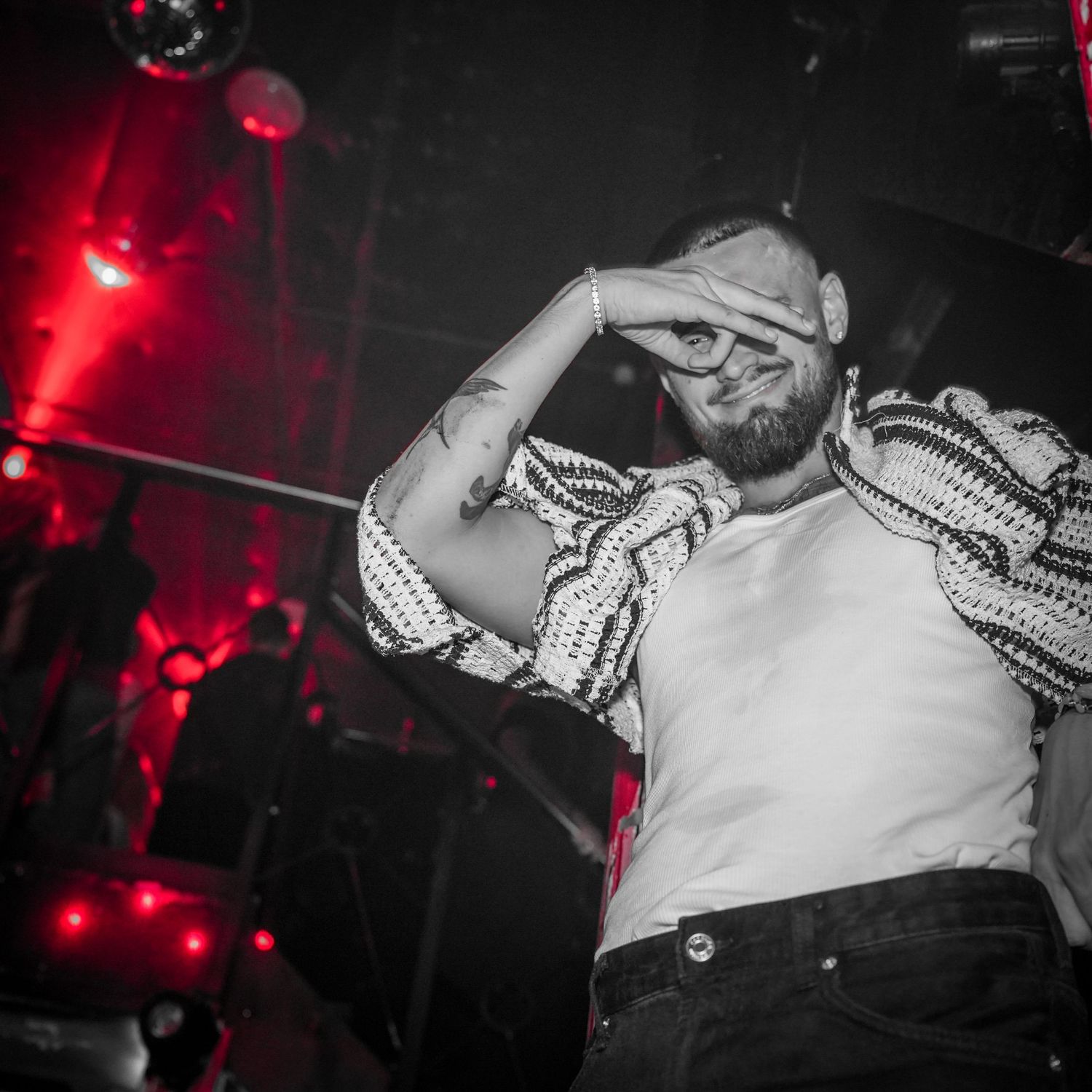 A performer in a white shirt and dark pants gestures dramatically on stage under dramatic red lighting at a concert venue.