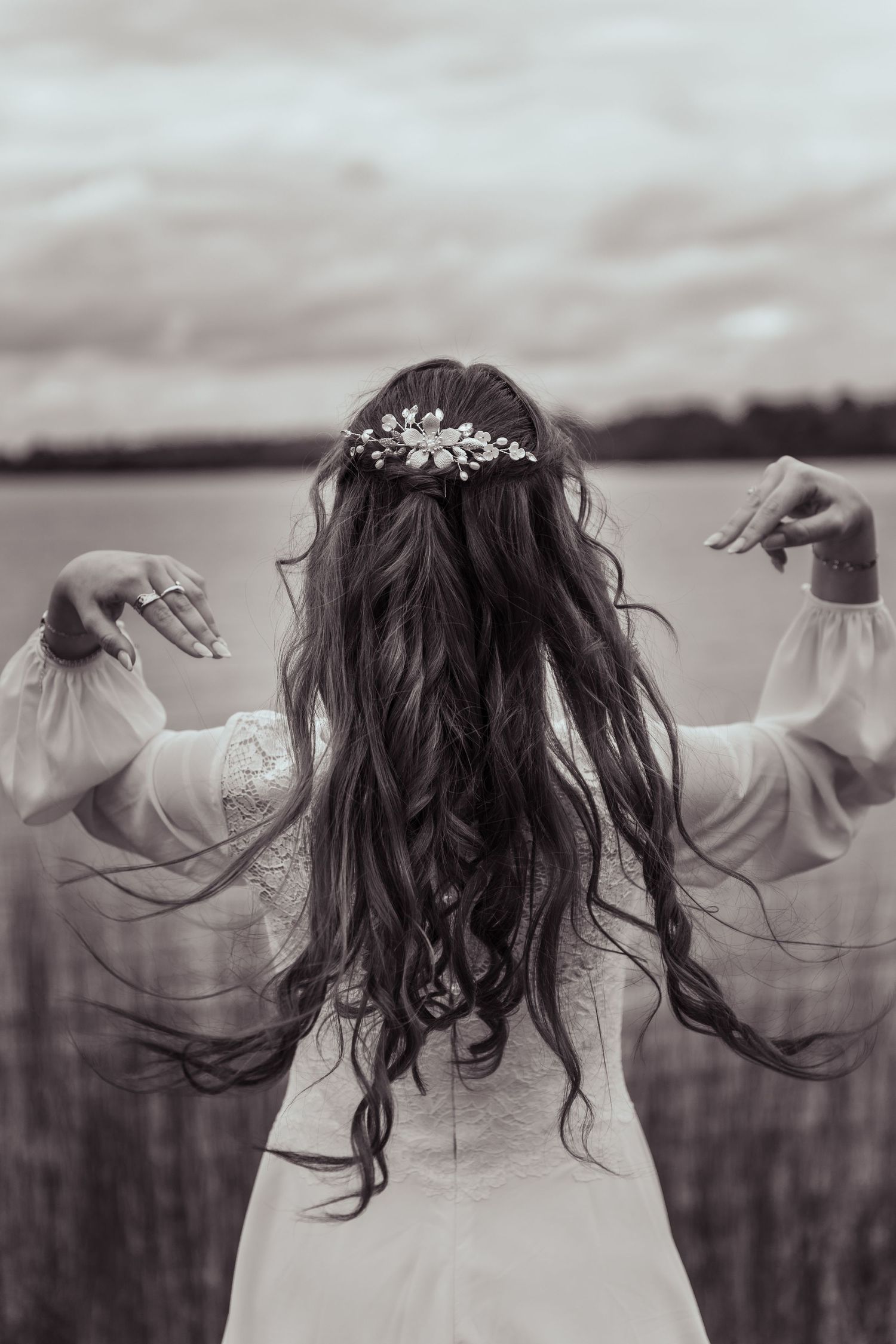 Back view of bride in flowing sleeve wedding dress with long dark hair and floral hair piece by a lake.