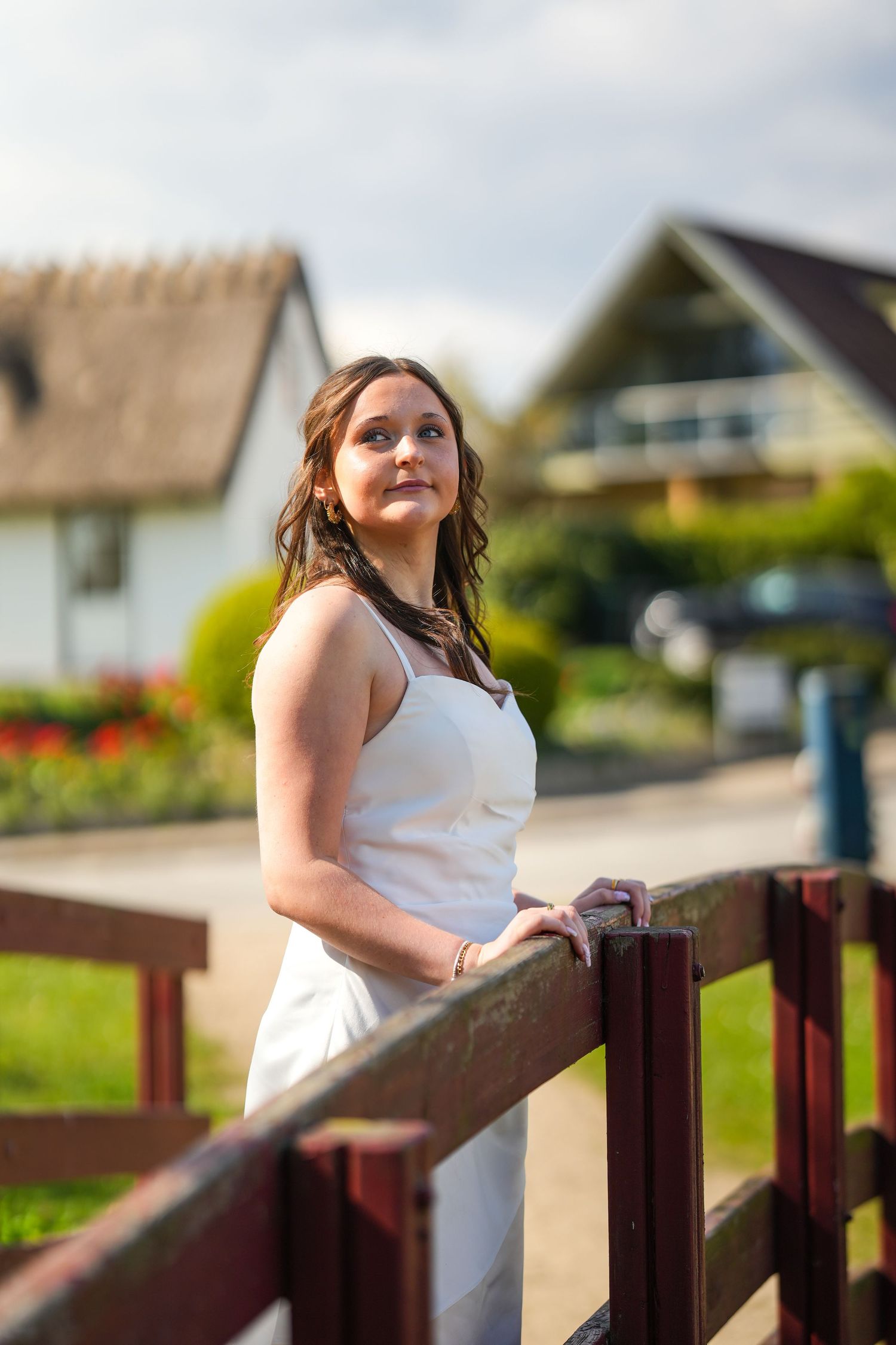 Person in white sleeveless dress leaning on wooden railing with houses and garden in the countryside background.