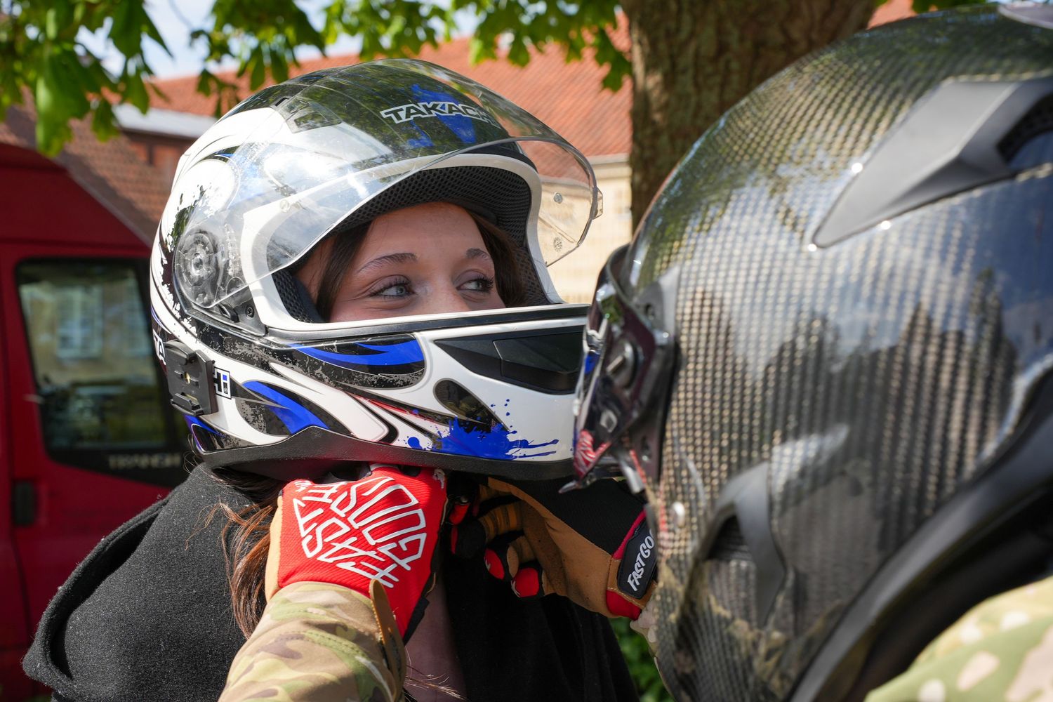 Two motorcycle helmets, one white with blue accents and one carbon fiber, being compared outdoors under green foliage.