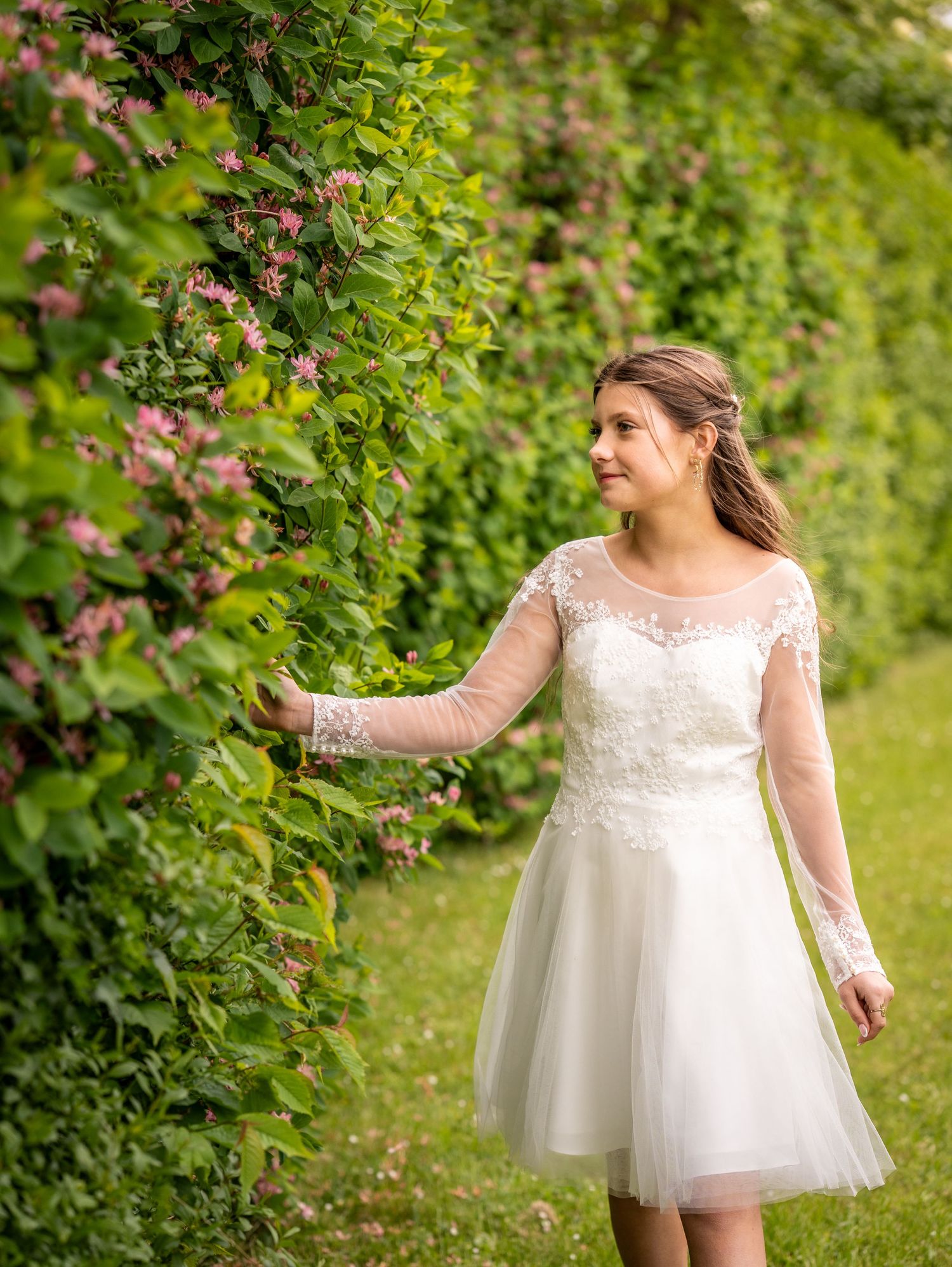 A bride in a short lace and tulle wedding dress reaches toward flowering vines in a lush garden setting.