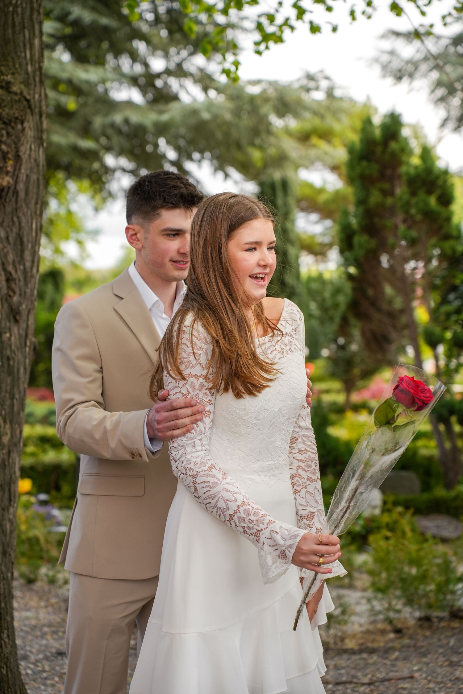 Newlywed couple in garden setting with bride in lace dress holding red roses and groom in tan suit.