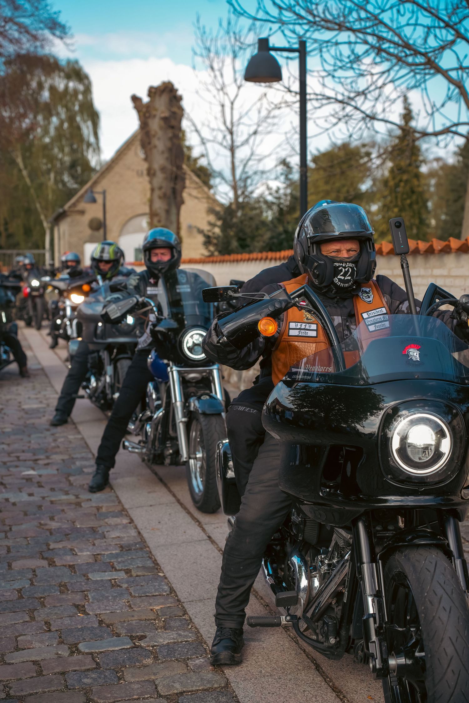 Motorcyclists in black gear lined up on a brick sidewalk with their bikes on an autumn day near residential buildings.