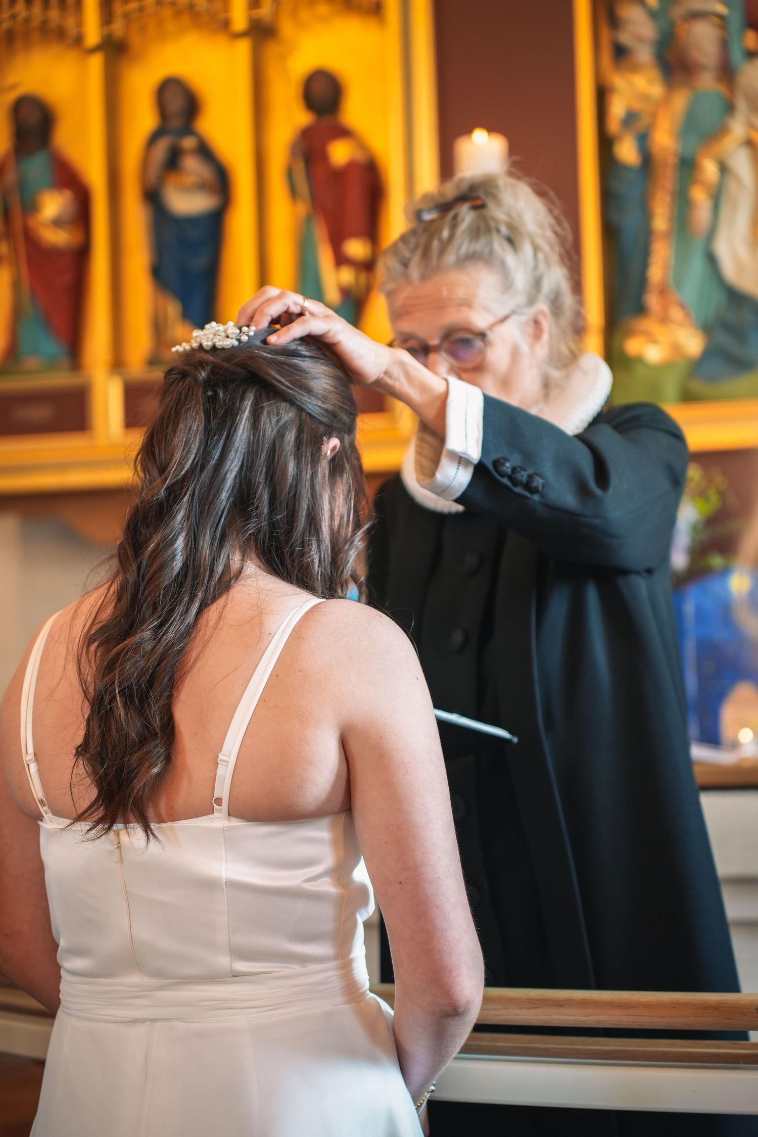 In a church with stained glass backdrop, a person in a black robe places a crown or tiara on someone wearing white dress.