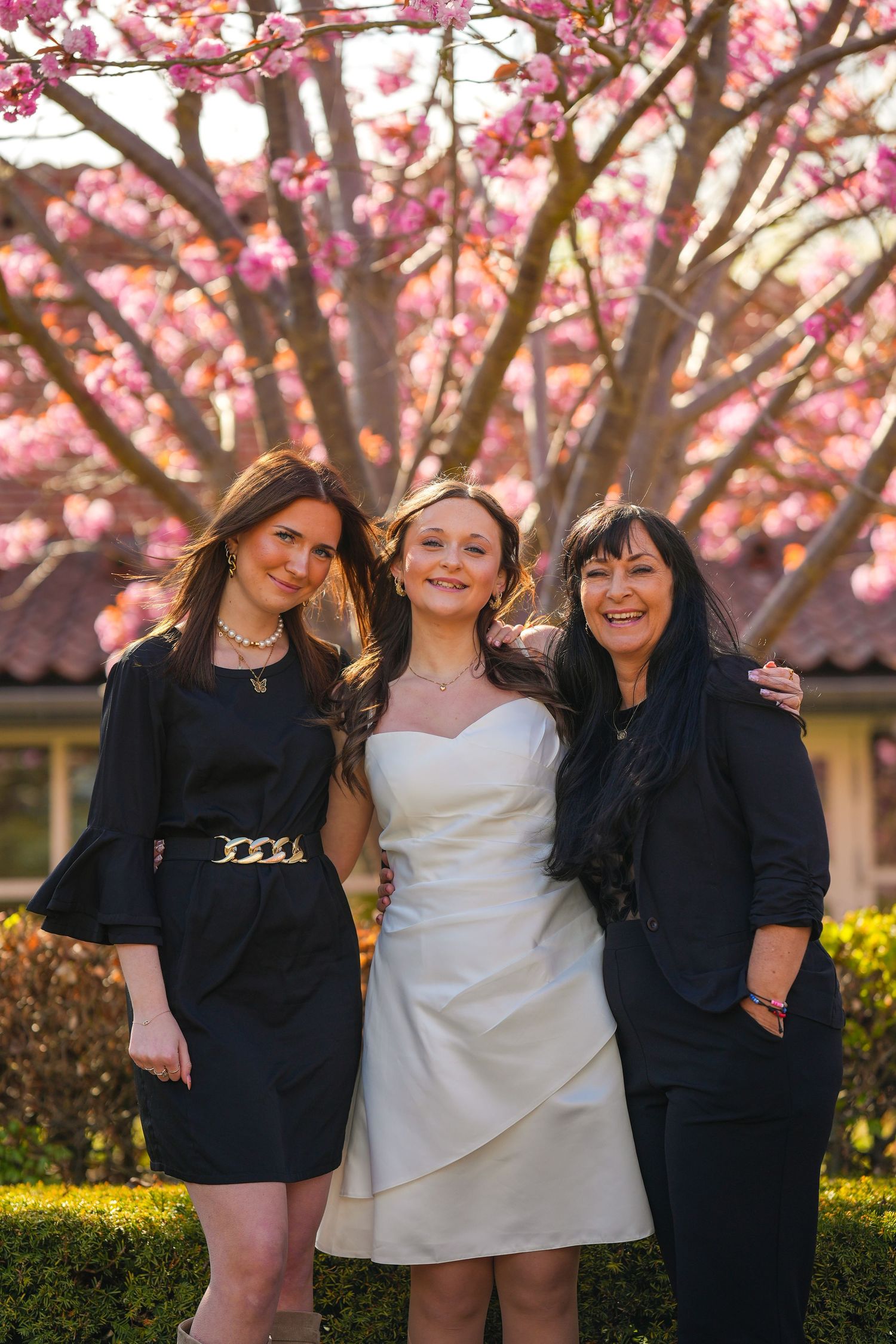 Three women pose together in front of blooming pink cherry blossoms on a sunny spring day.
