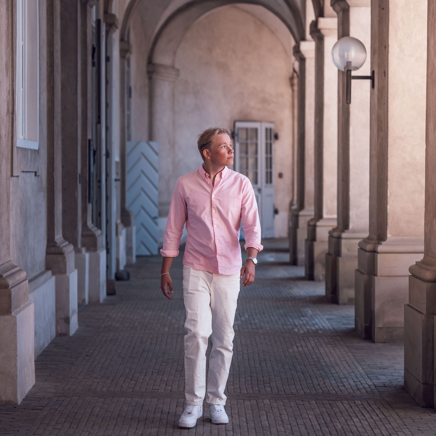 Person walking under ornate stone archway corridor wearing pink button-down shirt and white pants.