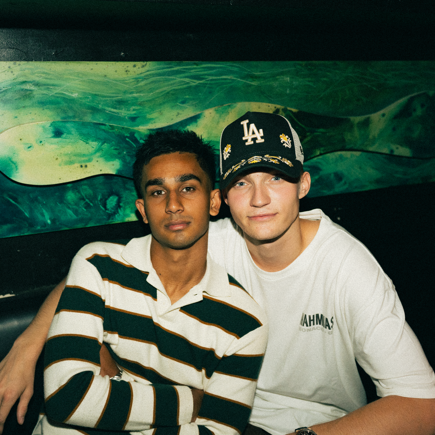 Two friends pose together in front of a green and yellow abstract background while wearing casual clothing.
