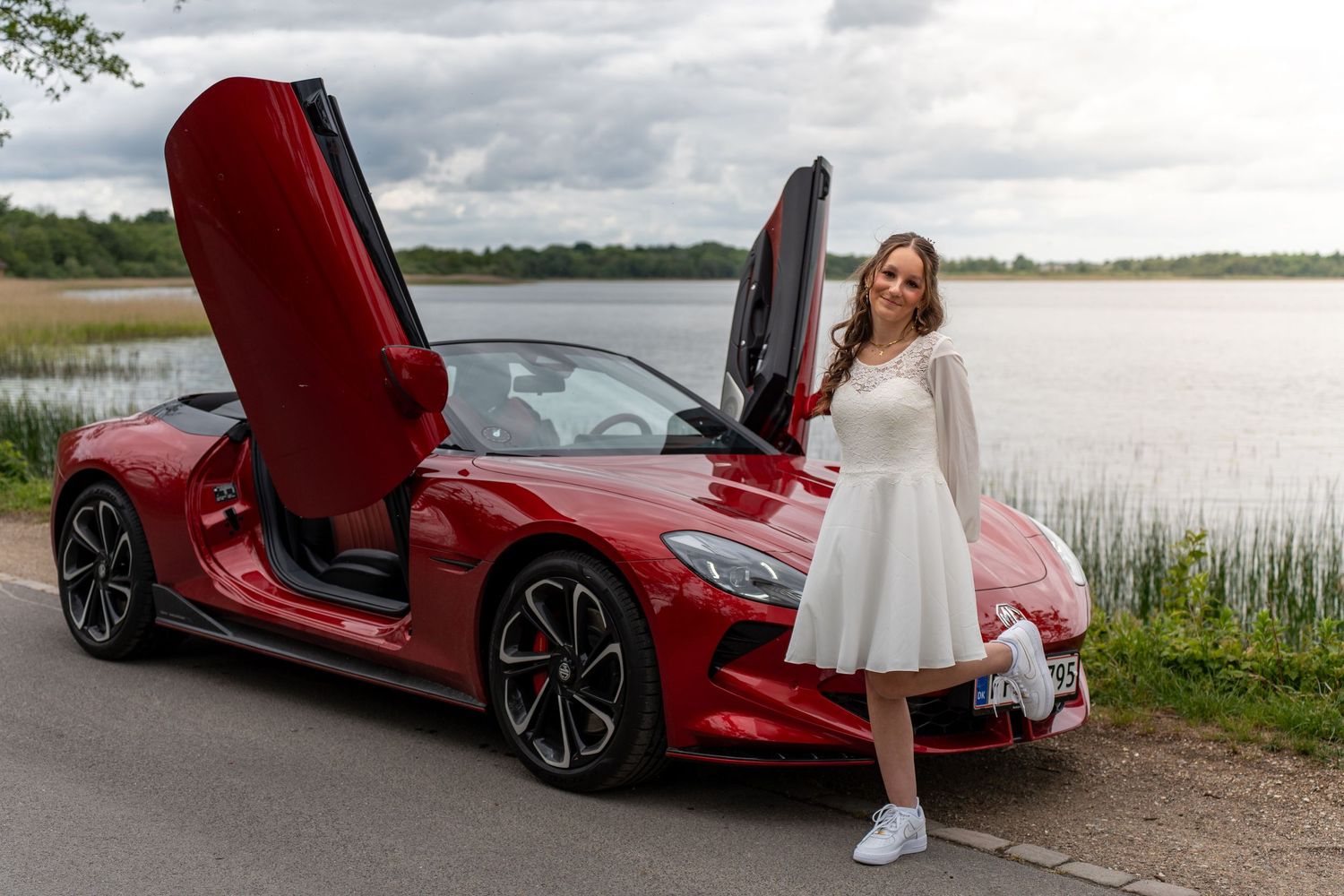 A glamorous red McLaren sports car with butterfly doors parked by a scenic lake under cloudy skies.