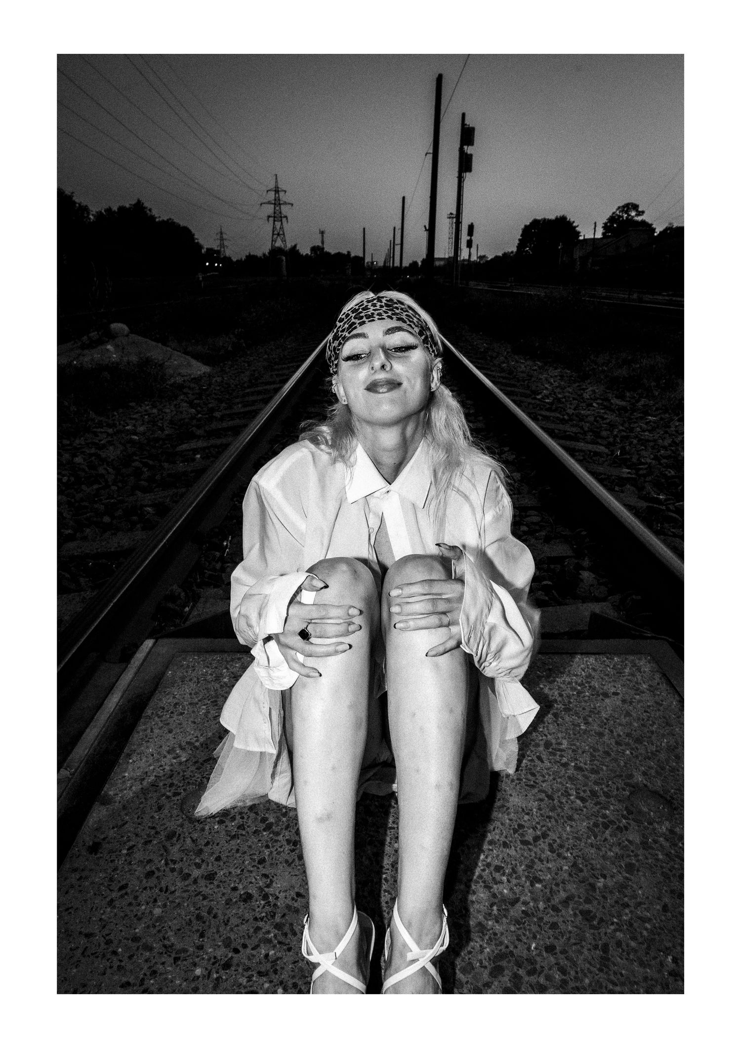 Black and white photo showing someone sitting on train tracks wearing a white shirt and shorts in a dramatic pose.