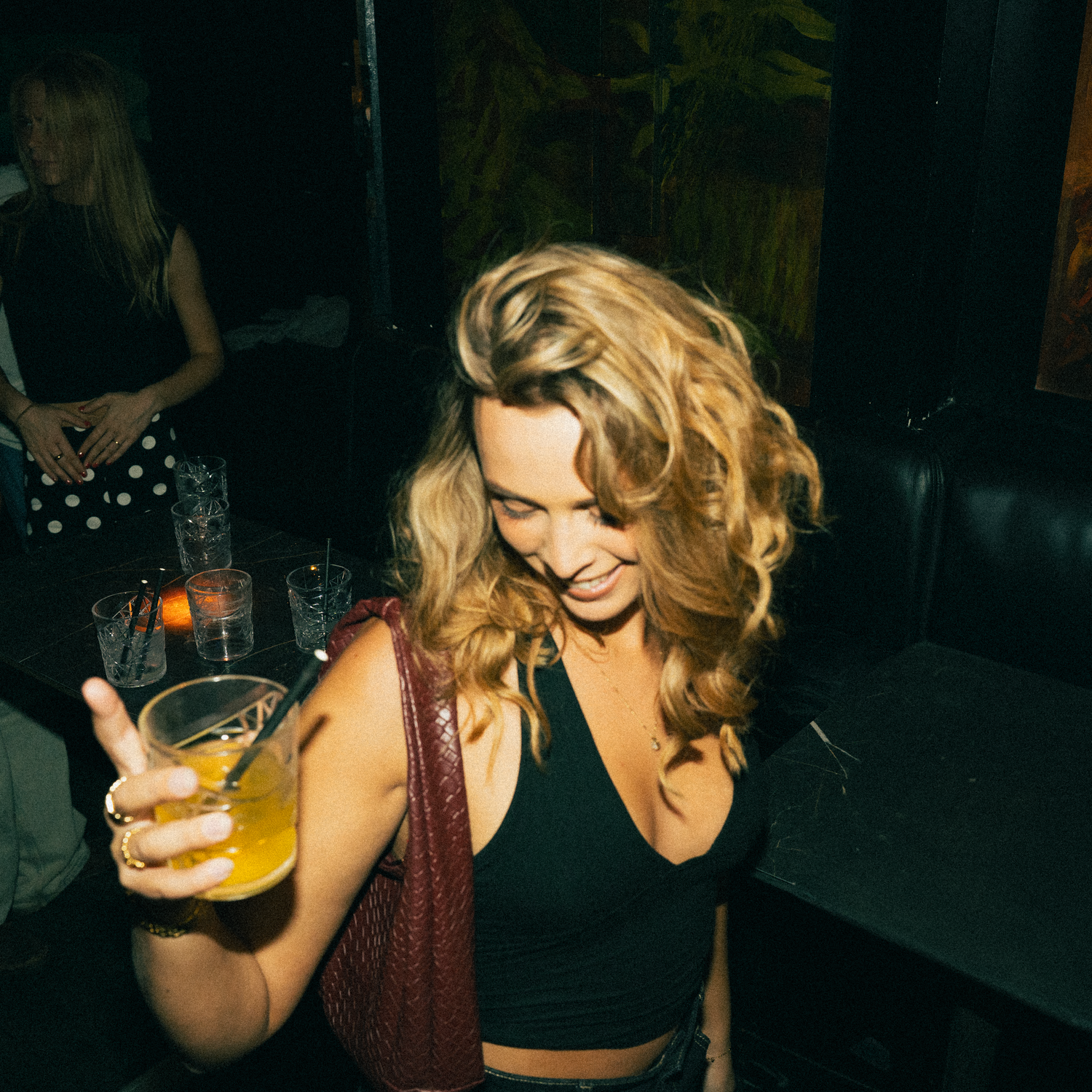 Person in dark crop top and vest holds orange cocktail at dimly lit nightclub while dancing.