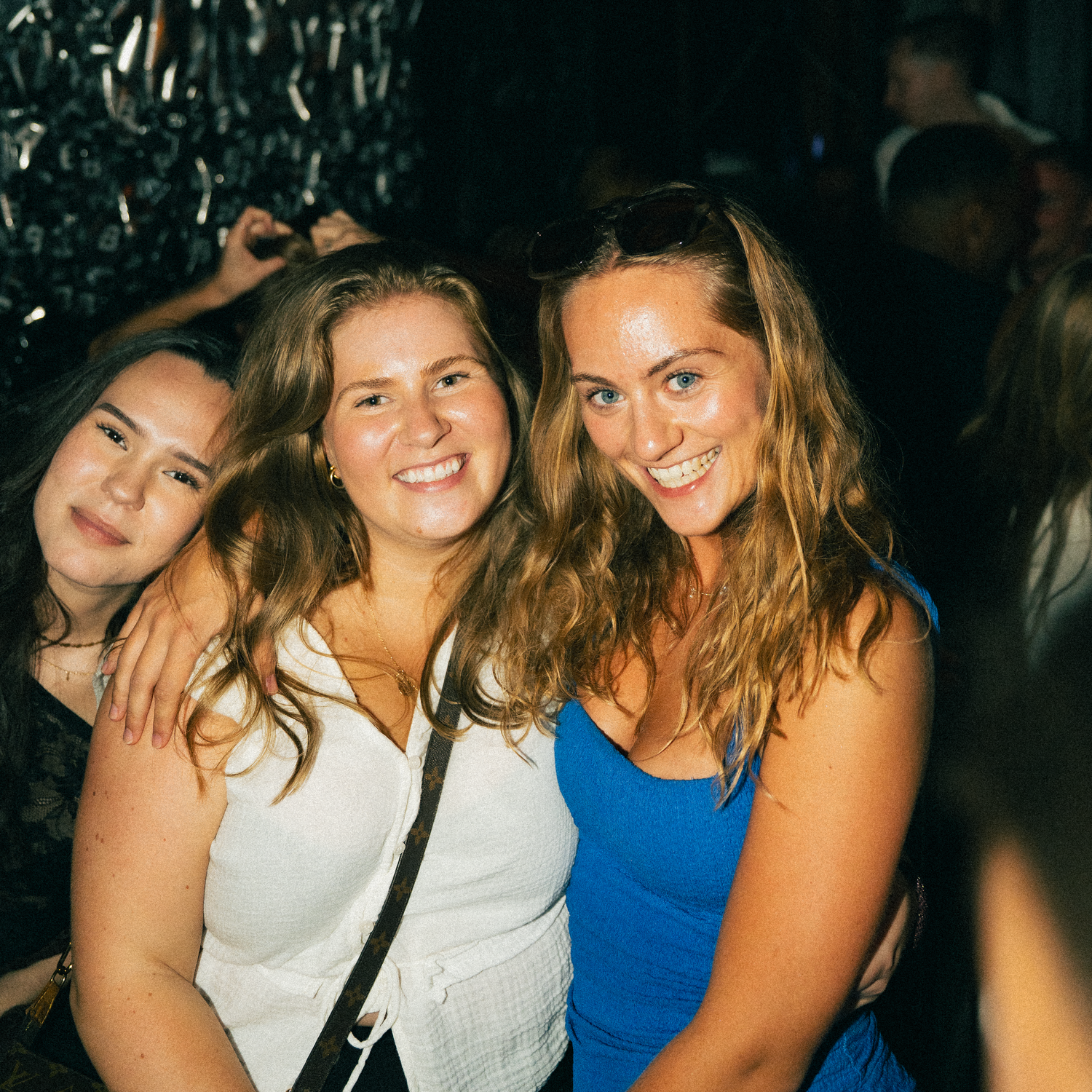 Three friends smile together at a nighttime social gathering, with one wearing blue and another in white.