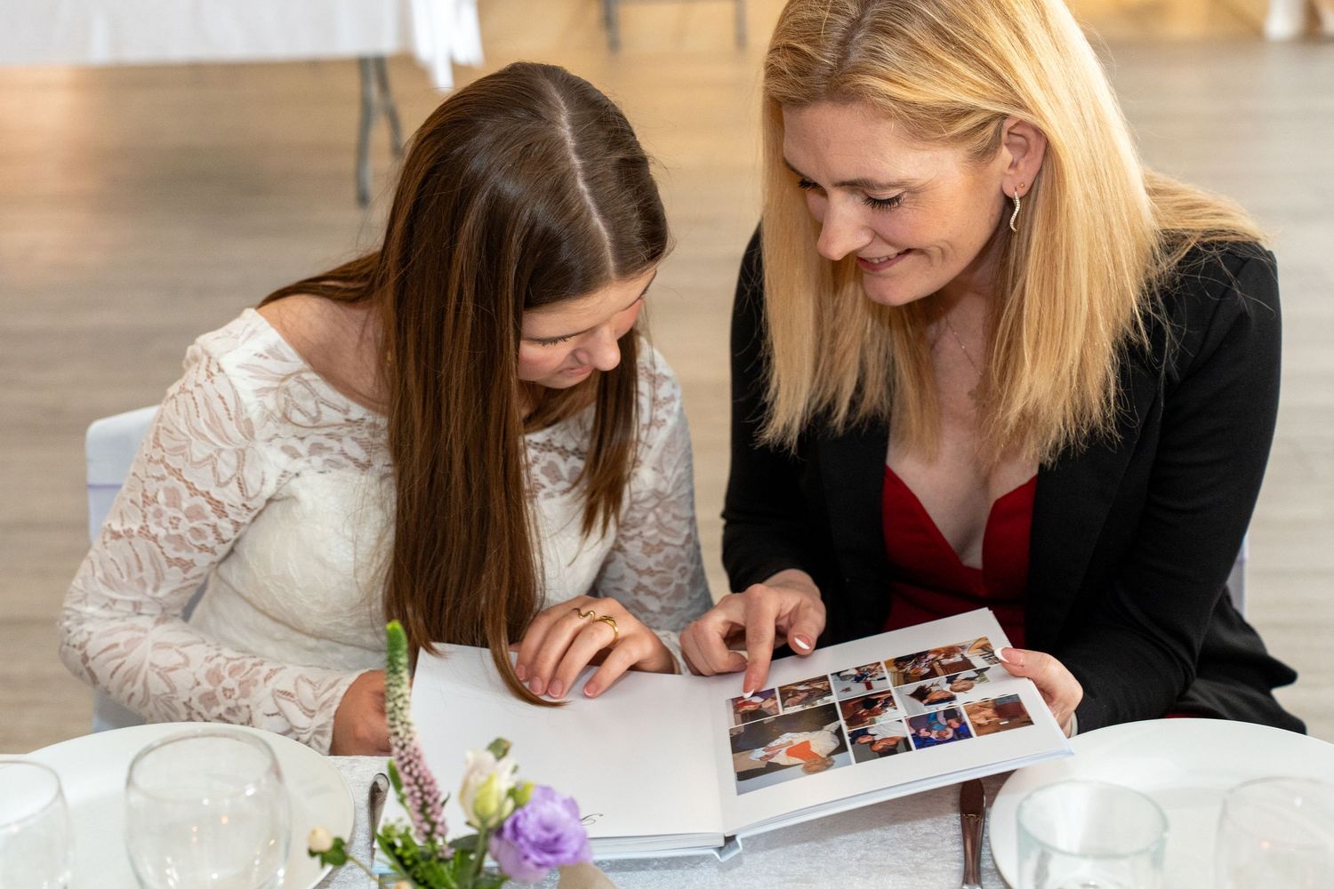 Two individuals look at a photo album together at a table decorated with a vase of purple flowers.