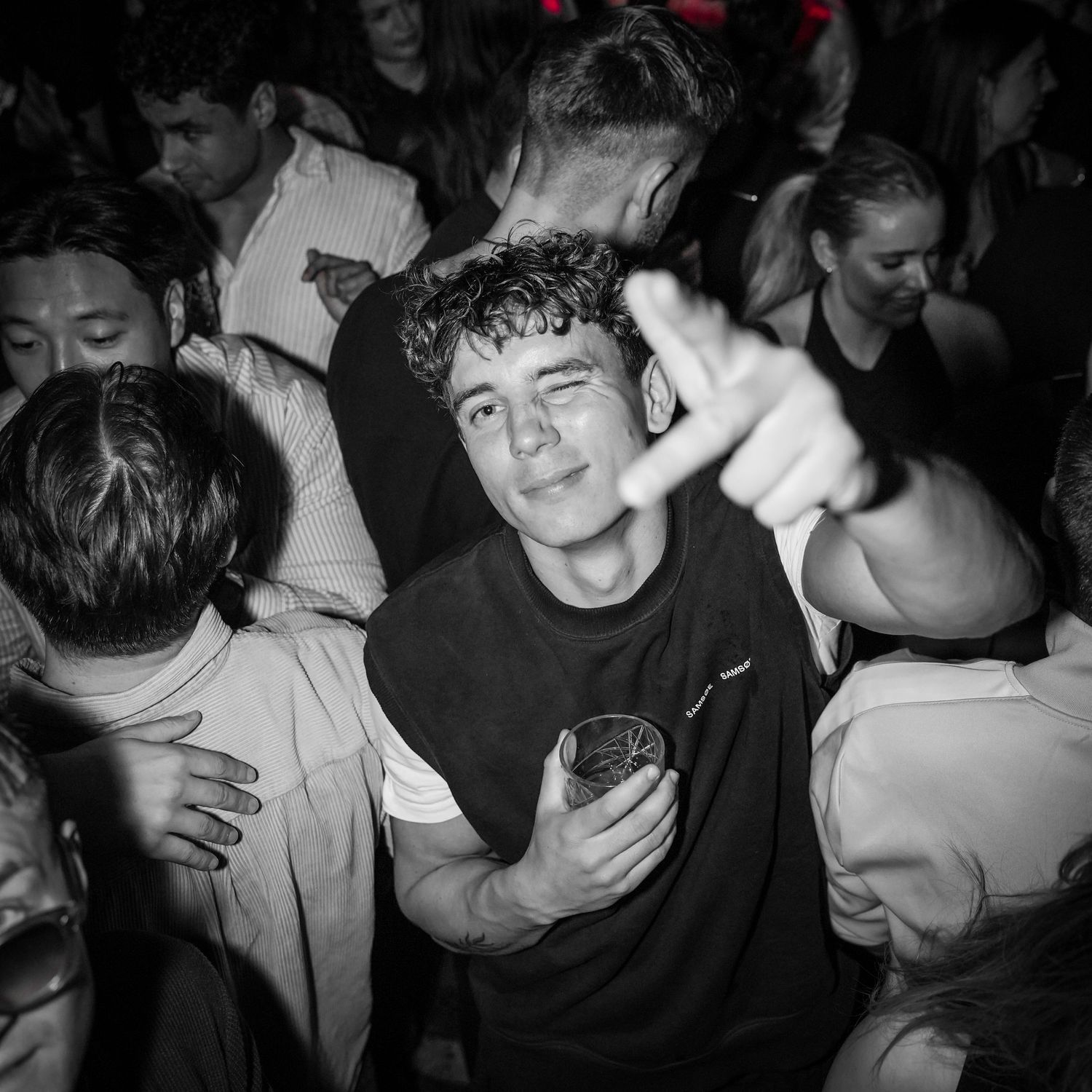 Black and white photo of a person dancing at a crowded nightclub while holding a drink in their hand.