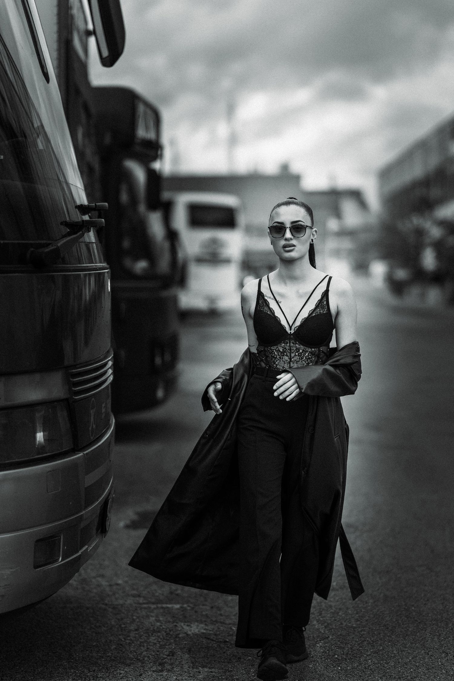 Artistic black and white fashion photo with model in sunglasses and long coat walking between parked buses on urban street.