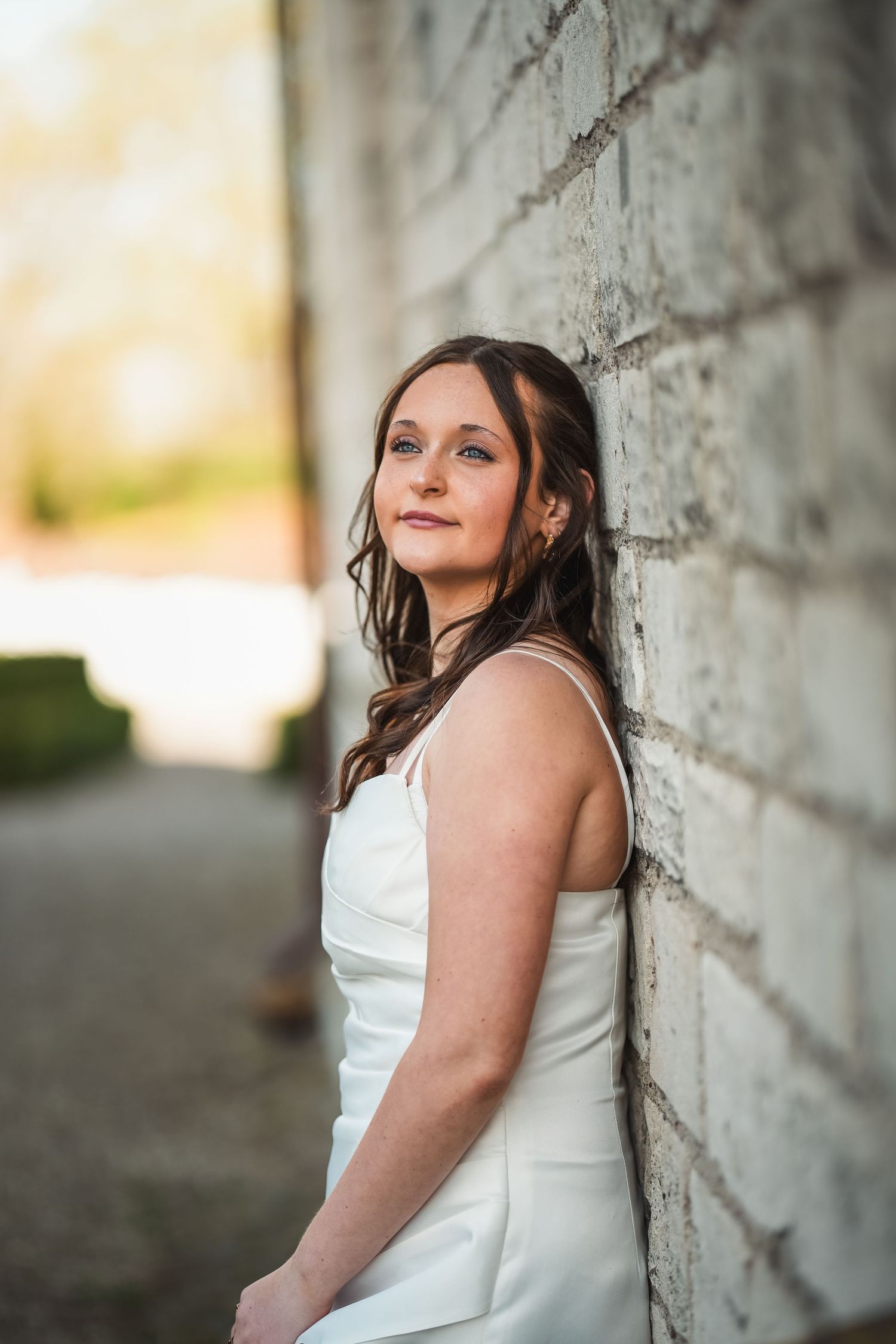 Person in a white dress poses against a textured gray wall in warm outdoor lighting creating an elegant portrait.