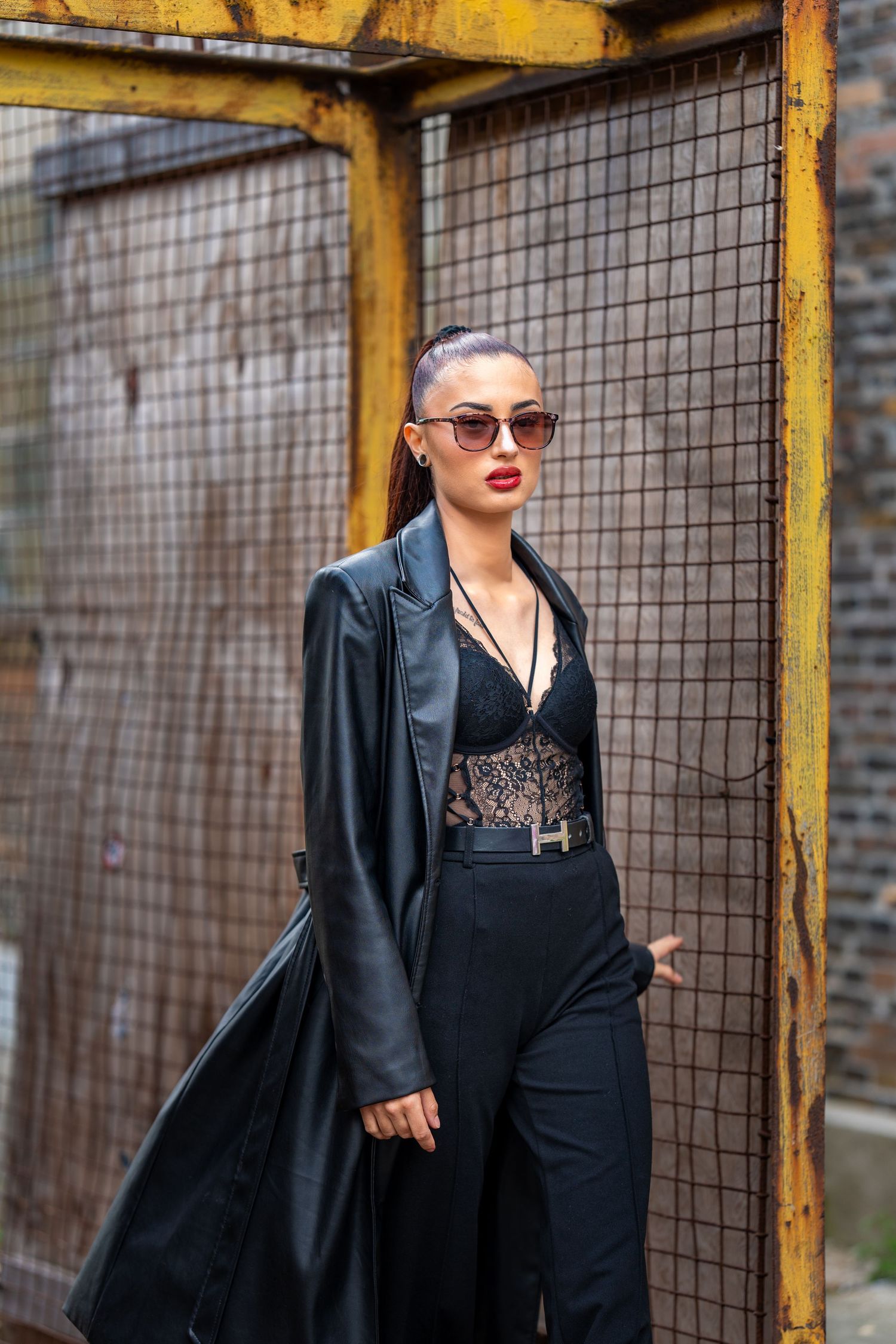 Fashionable gothic style outfit featuring a black blazer, lace top and sunglasses against an industrial backdrop.