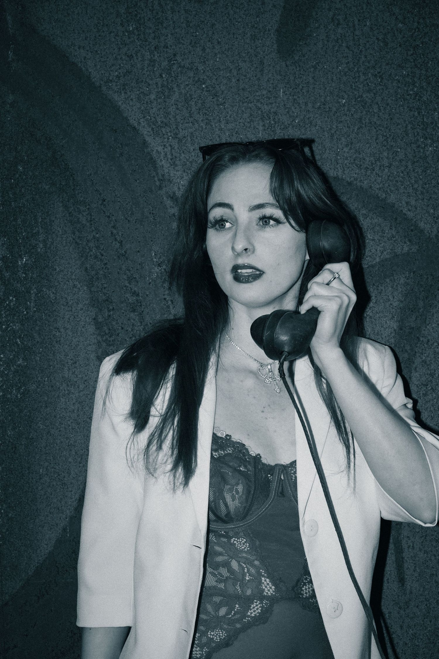 A woman in a white blazer and lace top poses with a vintage telephone in a moody black and white photo.