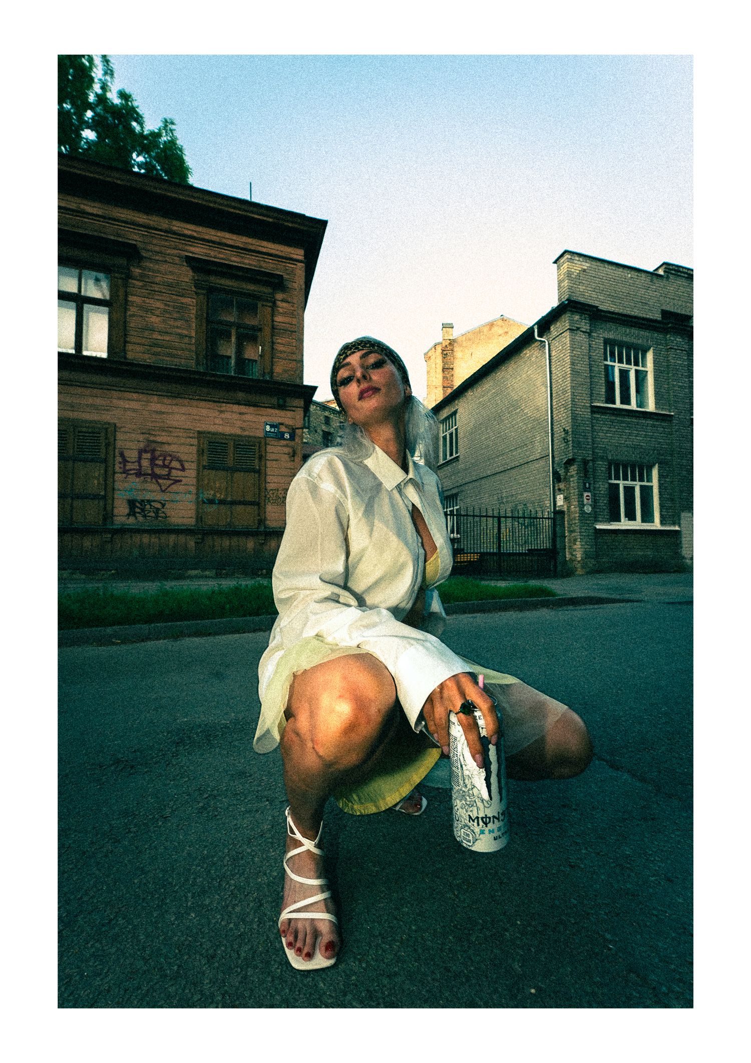 A person in a white shirt sits casually on an urban street corner with buildings in the background during dusk.