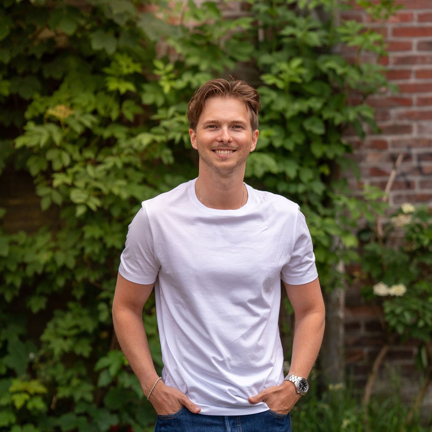 A person in a white t-shirt stands casually with hands in pockets against a brick wall with green ivy.