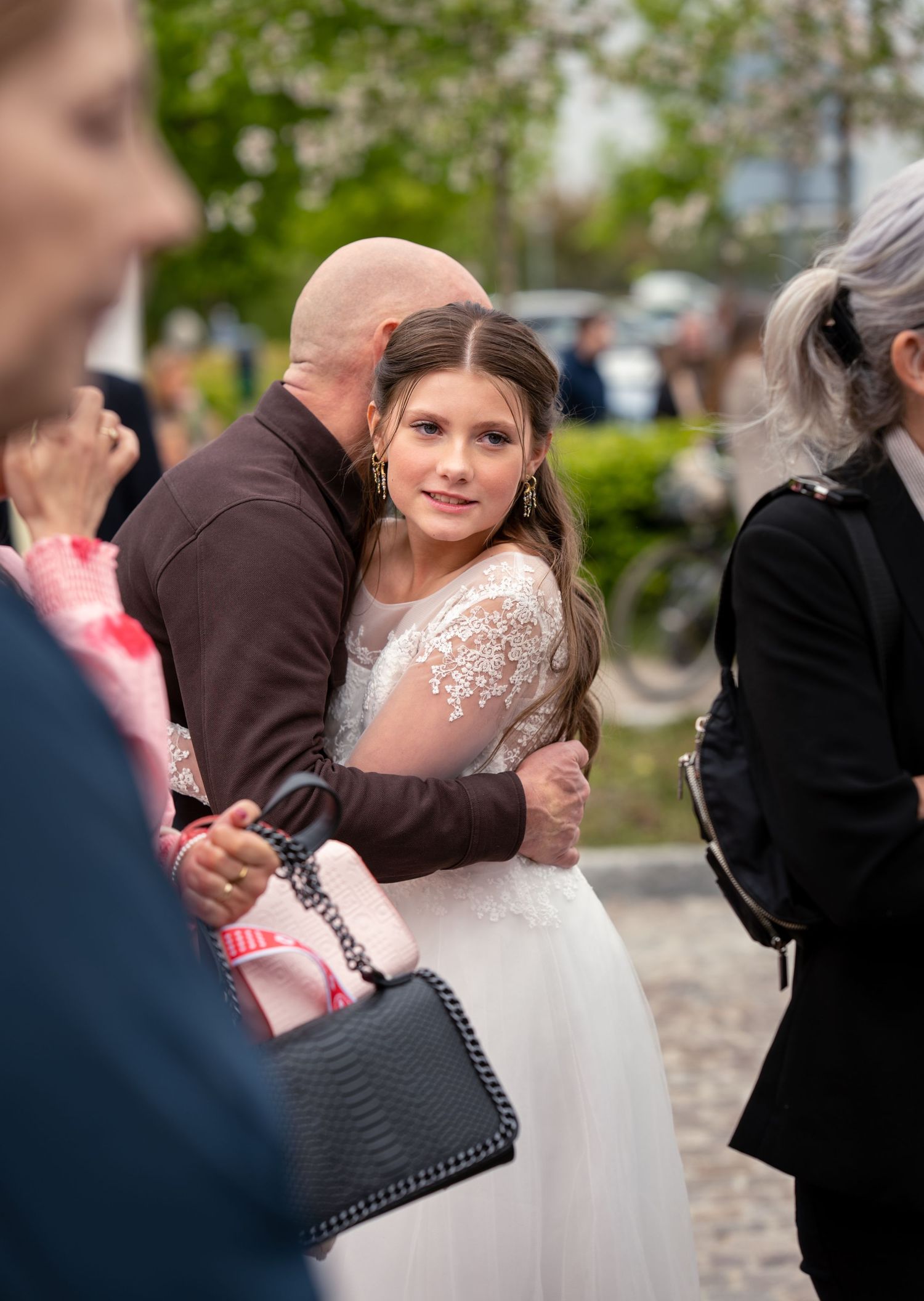 People gathering at an outdoor event as someone wearing a white lace wedding dress embraces another person.