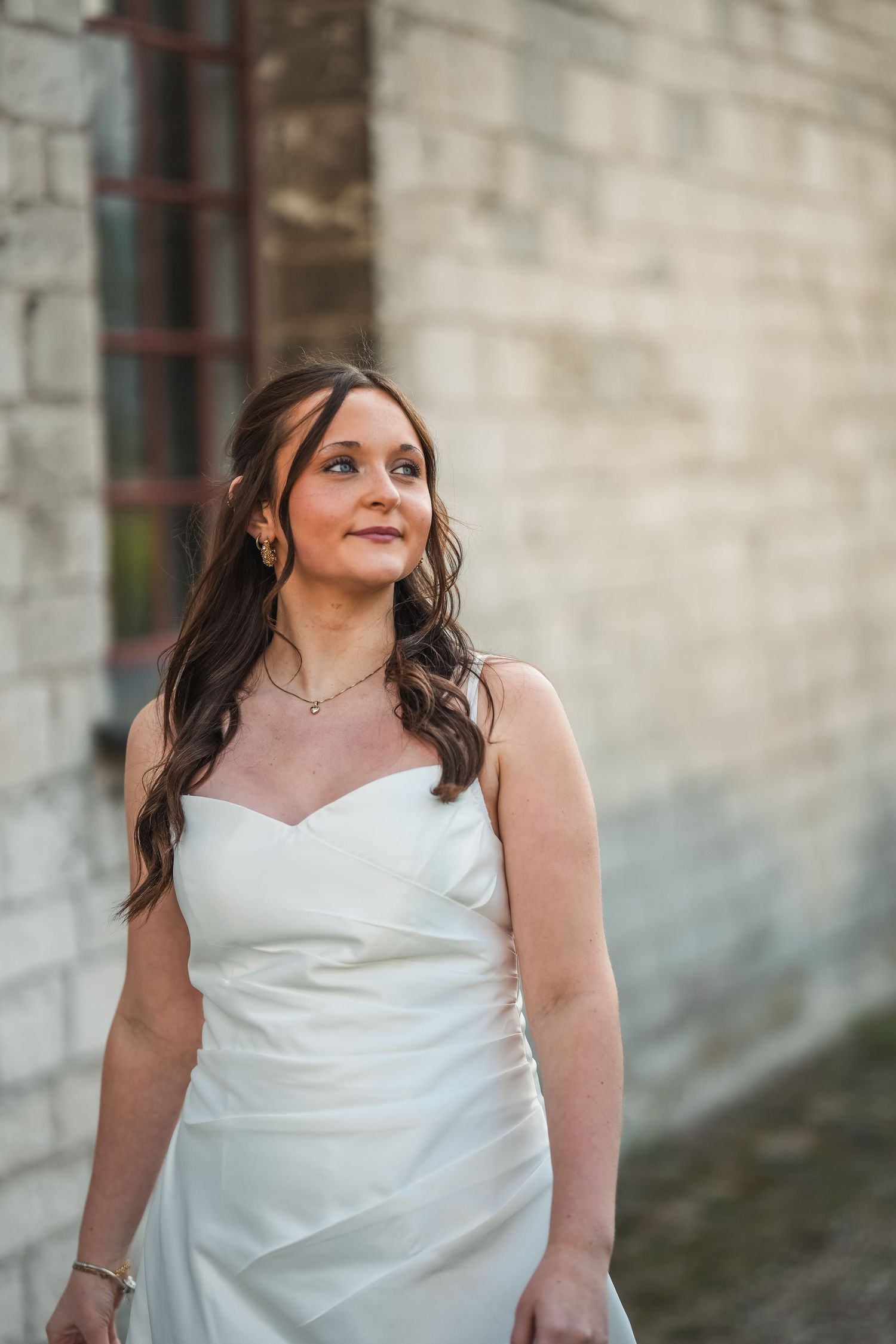 A bride in a strapless white wedding dress poses gracefully against a stone wall in soft lighting.