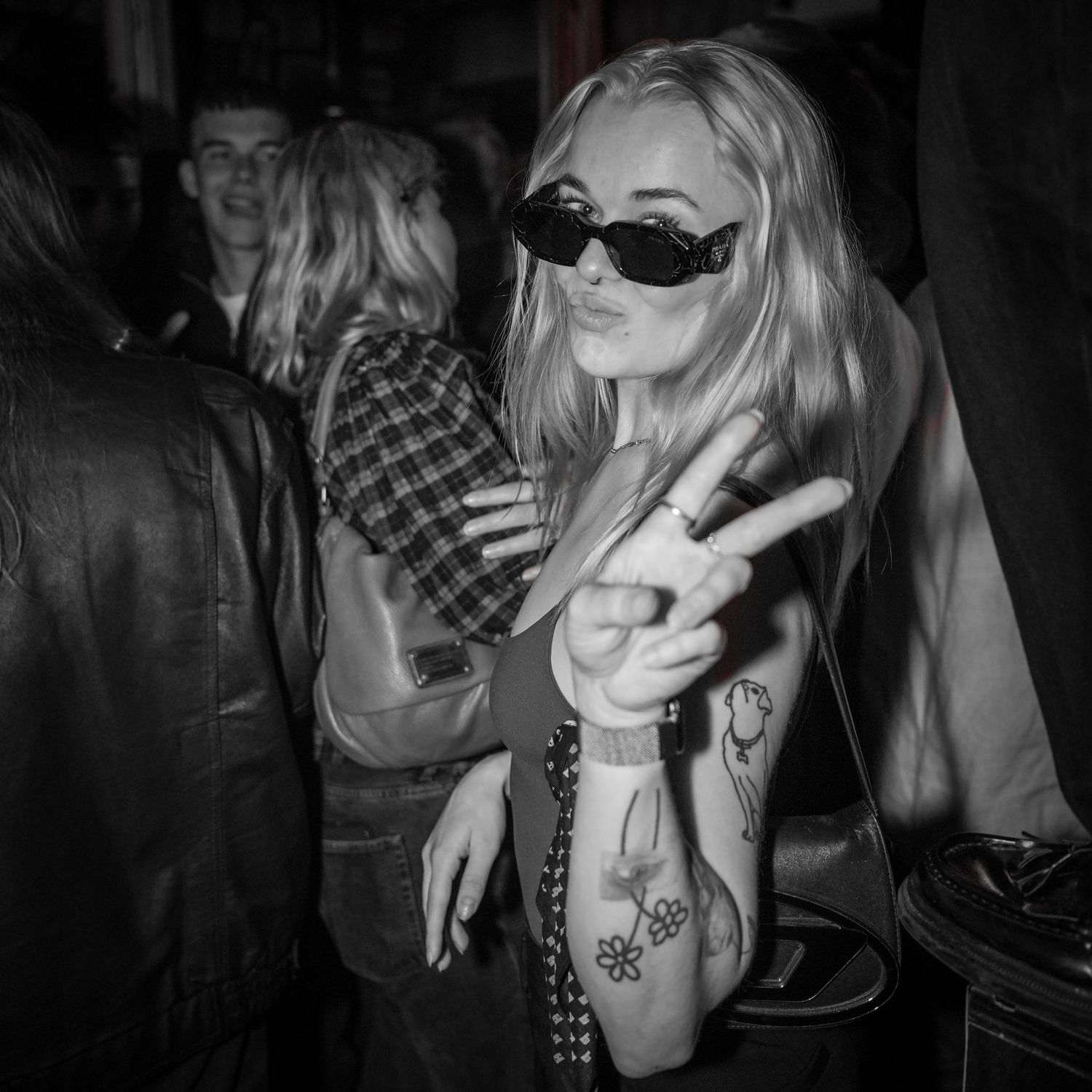 Black and white photo of person in leather jacket making peace sign while wearing sunglasses at a party.