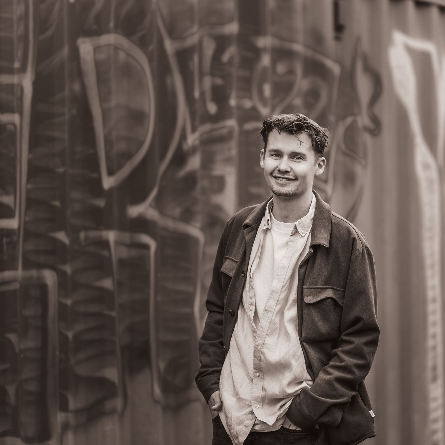Black and white portrait of a person smiling while standing against a graffiti wall wearing a jacket and backpack.