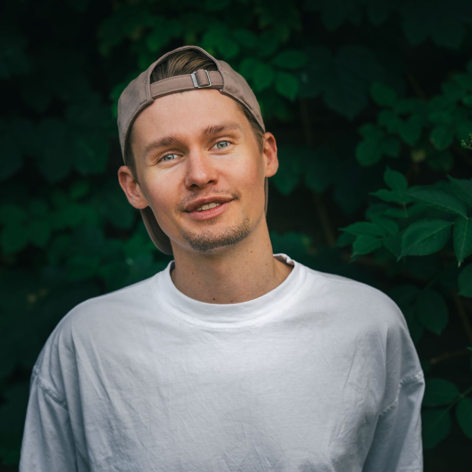 A casual portrait of someone wearing a backwards baseball cap and white t-shirt against lush green foliage background.