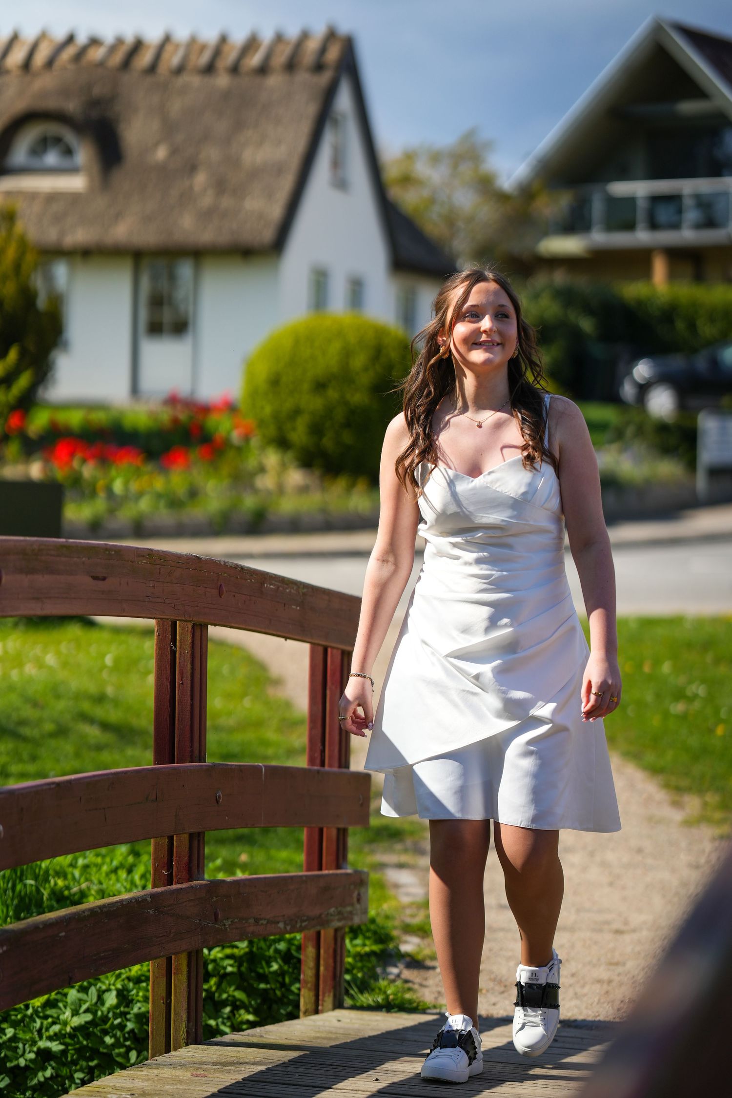 Person in white summer dress and sneakers walks across wooden bridge in front of charming cottage with thatched roof.