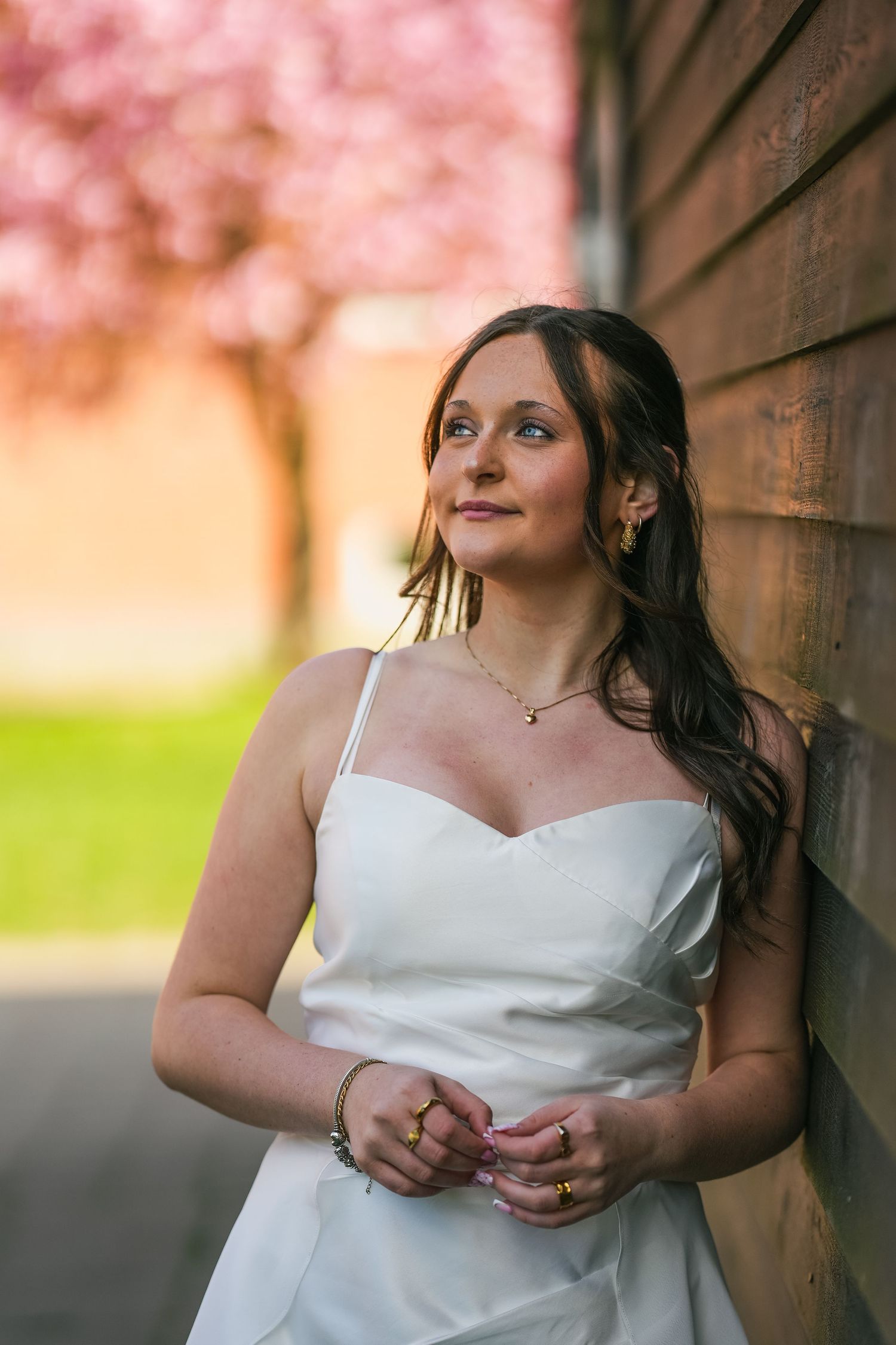 A bride in a classic white dress leans against a wall with pink cherry blossoms in the background during golden hour.