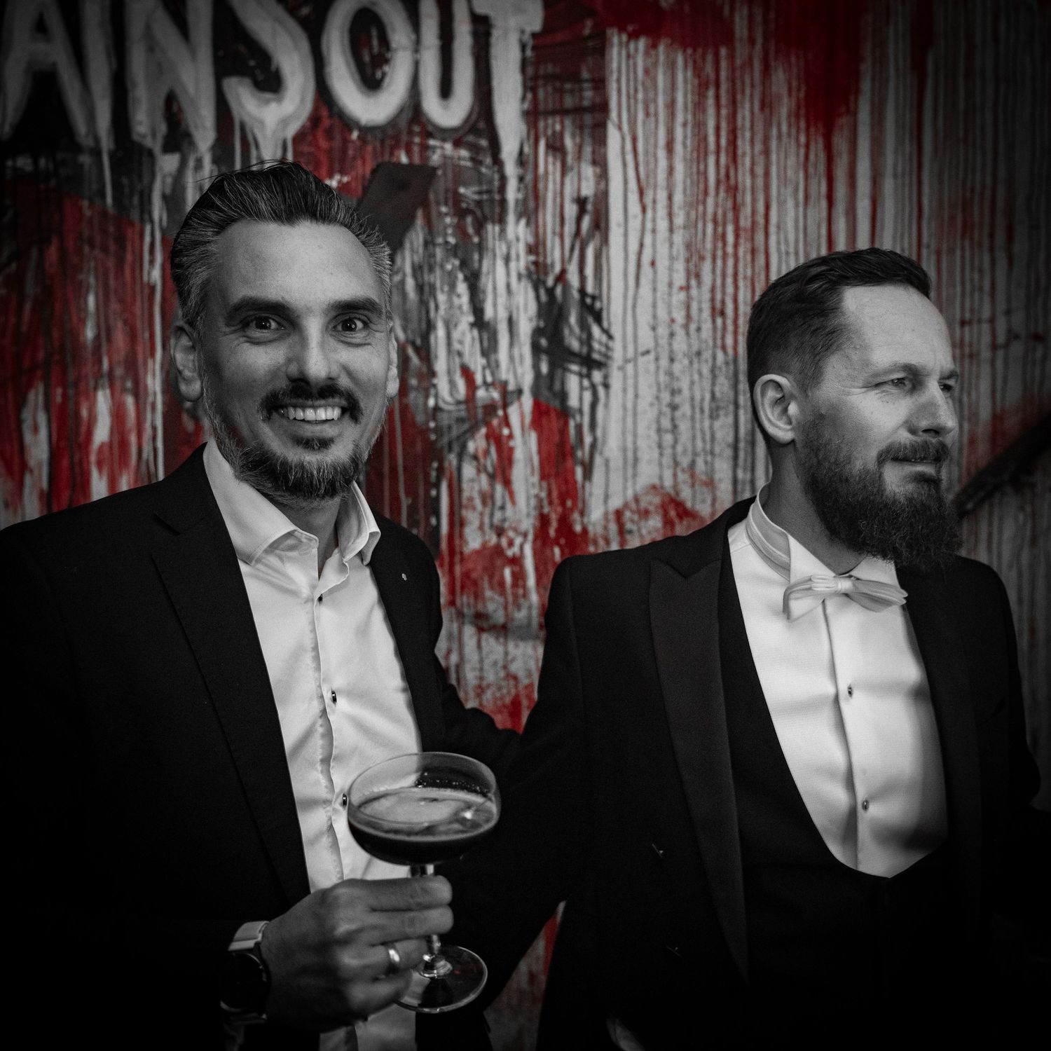 To velklædte gæster nyder drinks til et arrangement foran en dramatisk rød og hvid baggrund.