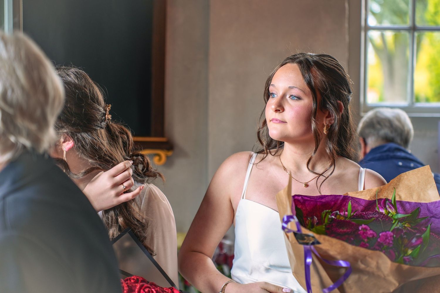 A person in a white satin dress holds a bouquet of purple flowers while others gather around at an indoor event.