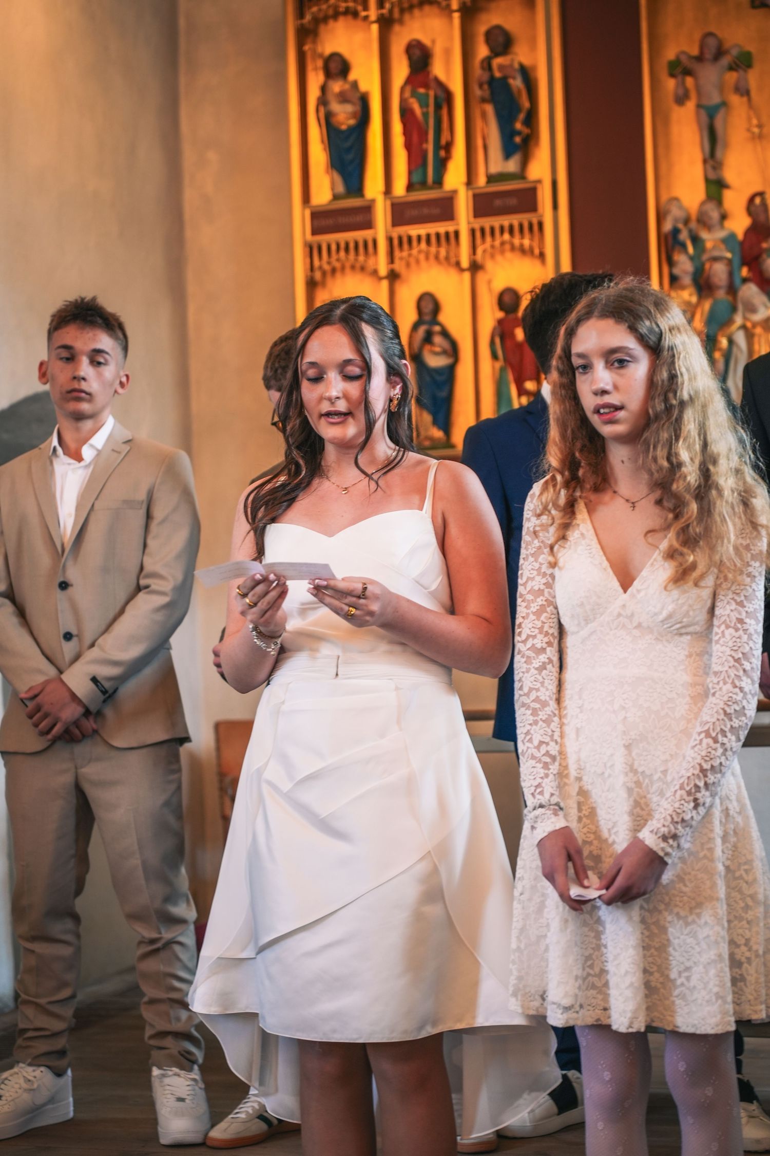 Religious ceremony with attendees in formal attire standing in front of ornate church wall with golden religious artwork.