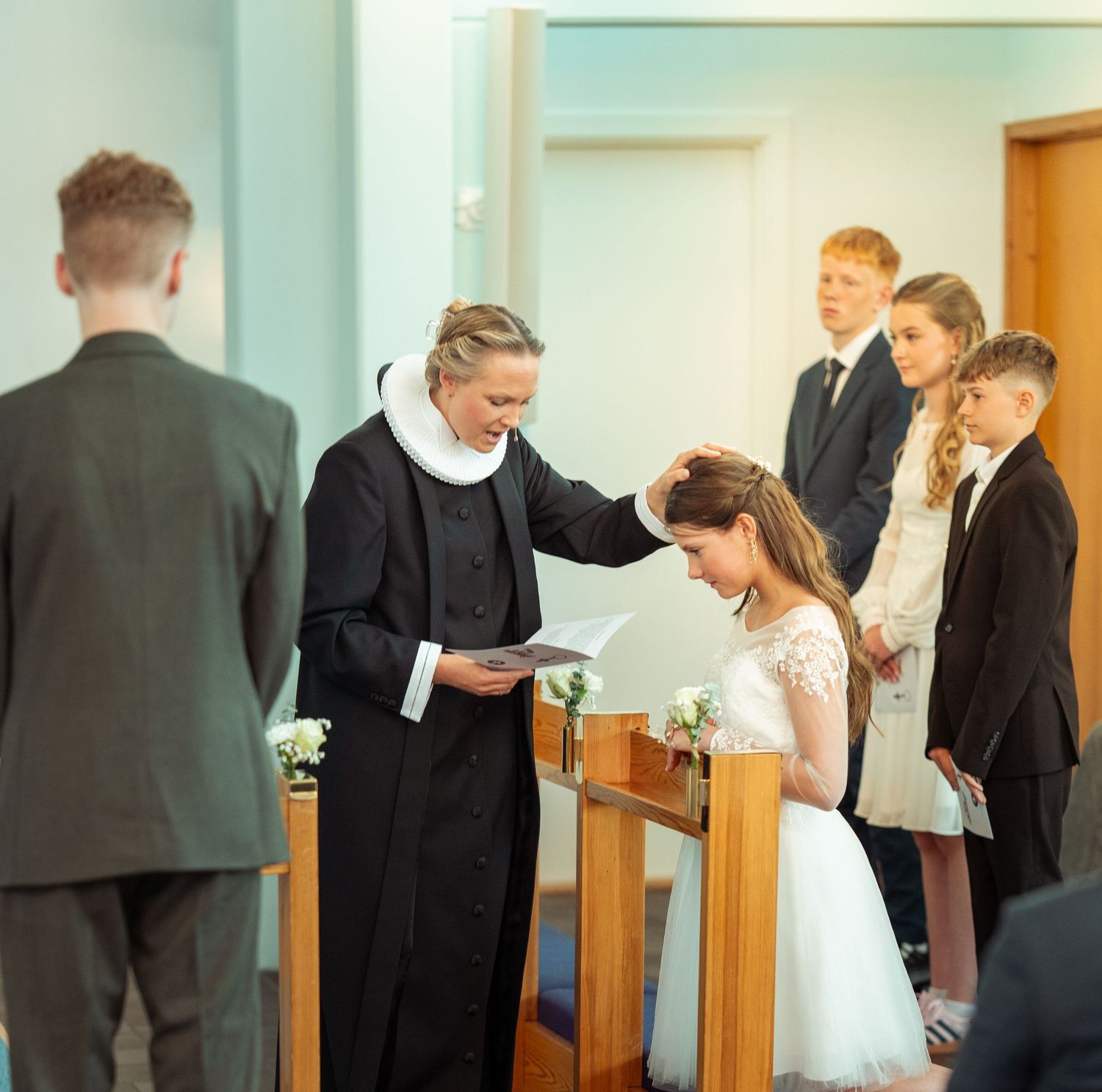 A minister blesses someone in a formal church ceremony while others look on in formal attire.