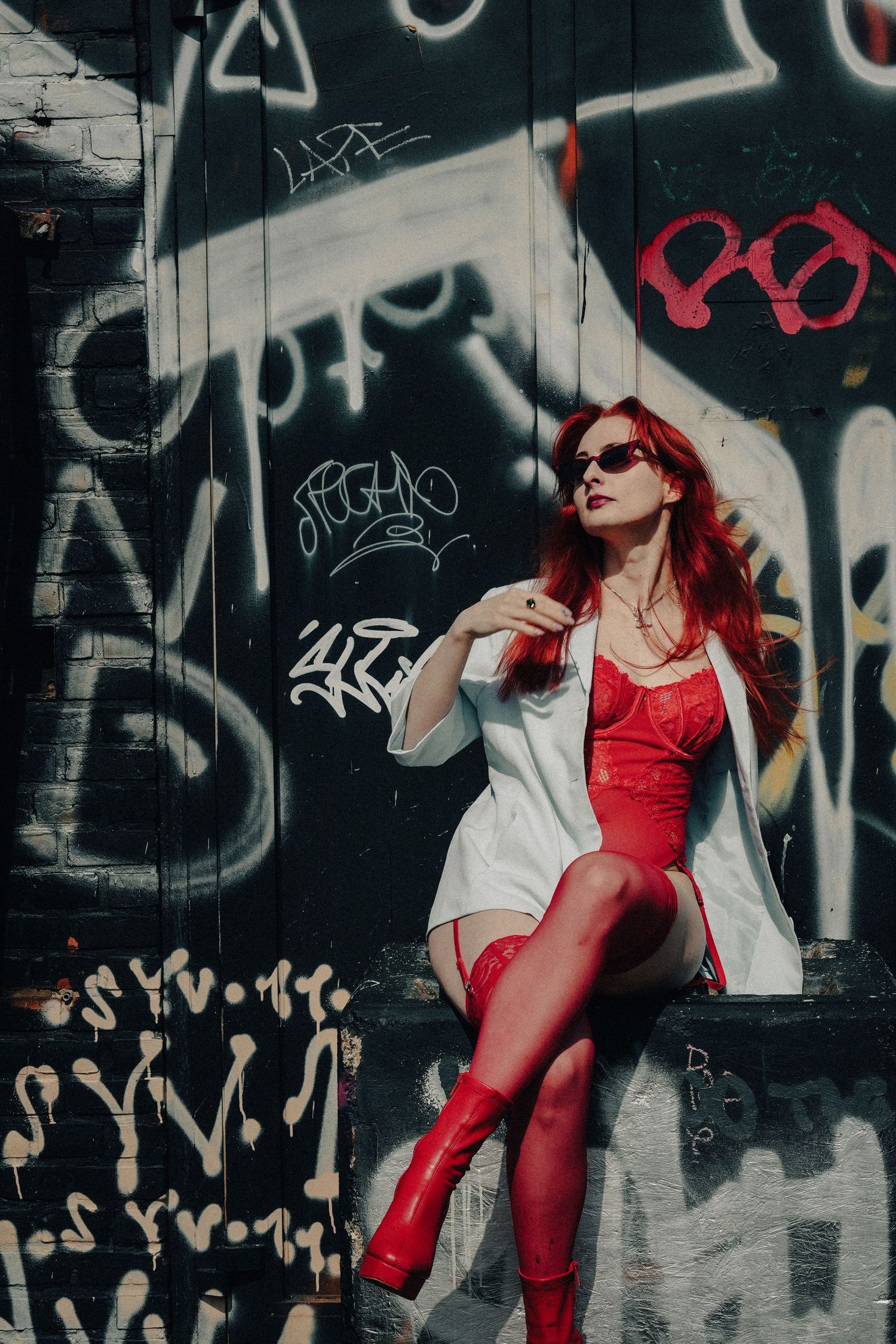 Fashion model in red swimsuit and white coat poses against graffiti wall wearing sunglasses.