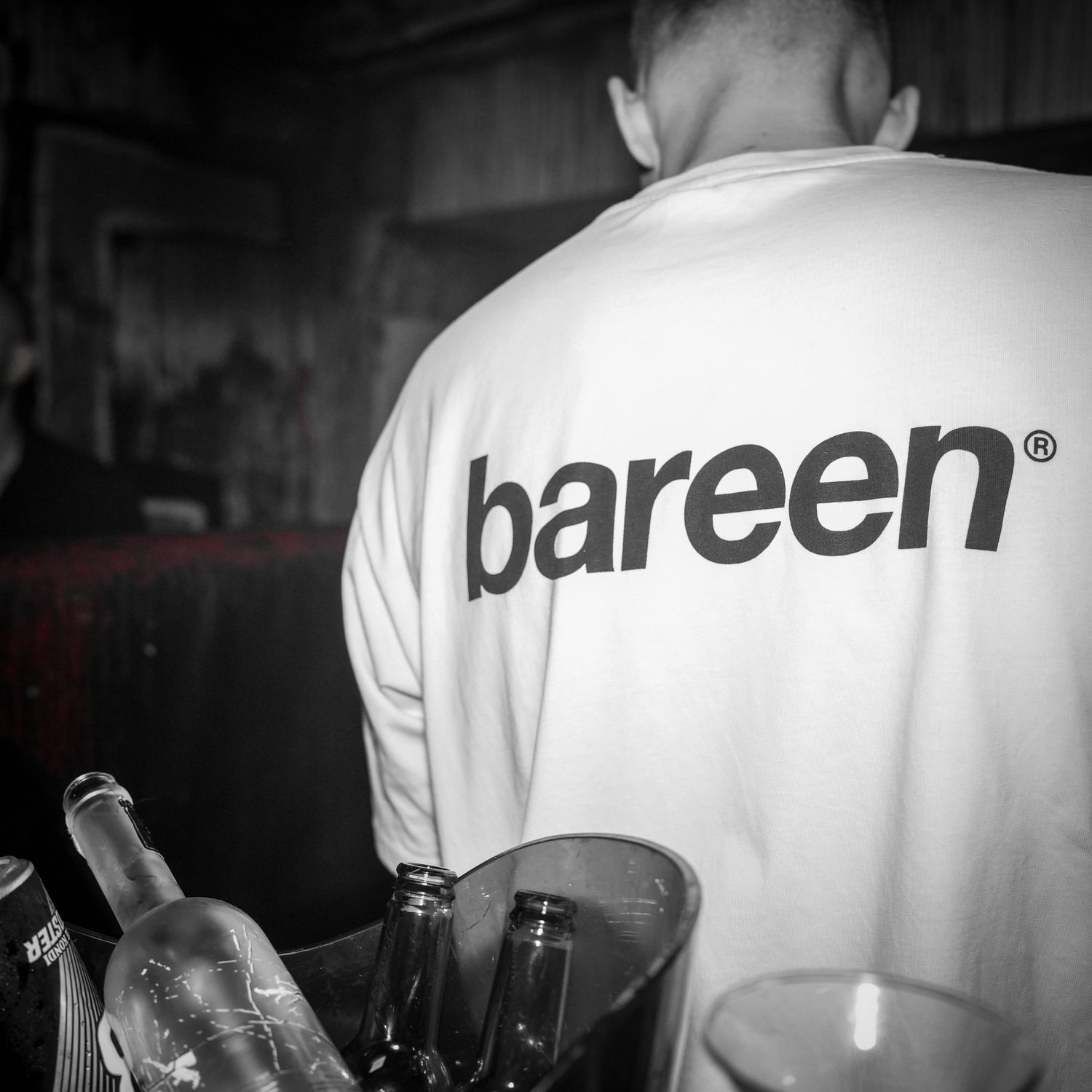A bartender wearing a white Bareen branded shirt shown from behind while attending to wine glasses and bottles.