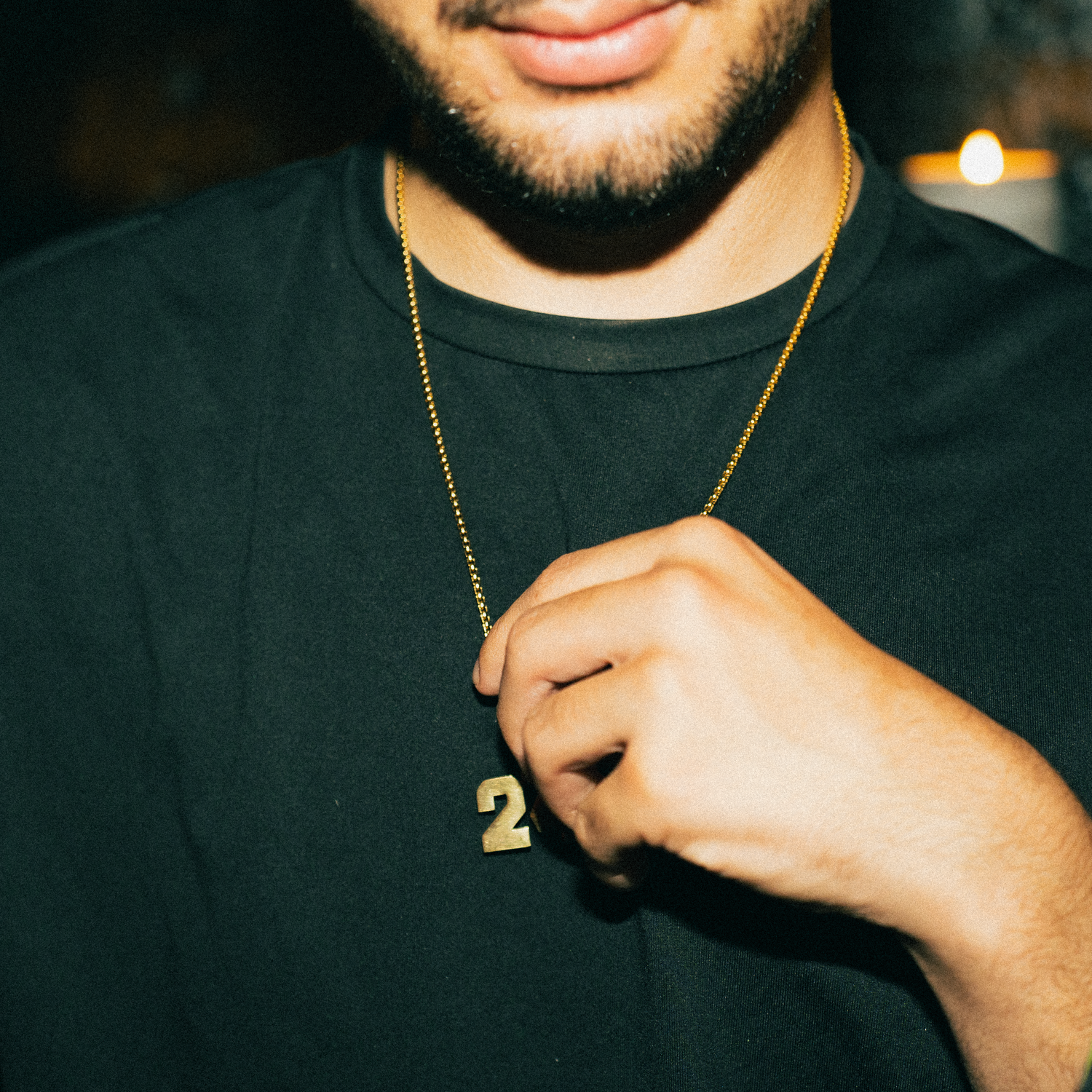 Close-up shot of someone wearing a black t-shirt and gold chain necklace with a number 2 pendant in dim lighting.