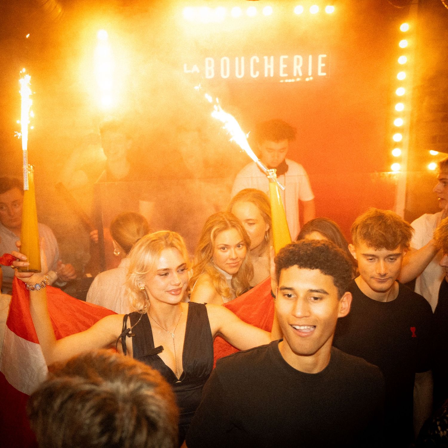 Folk danser tæt sammen på natklub med orange lys over dem.