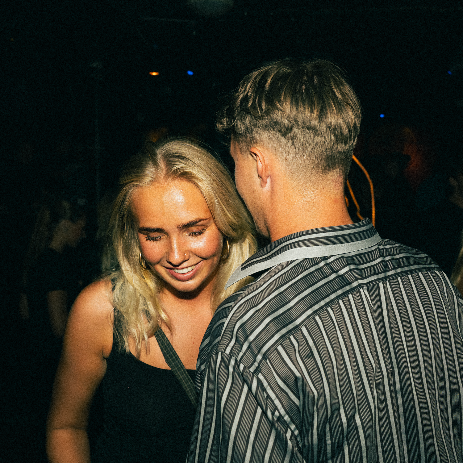 A couple shares an intimate dance moment in a dimly lit nightclub setting, with one person wearing a striped shirt.