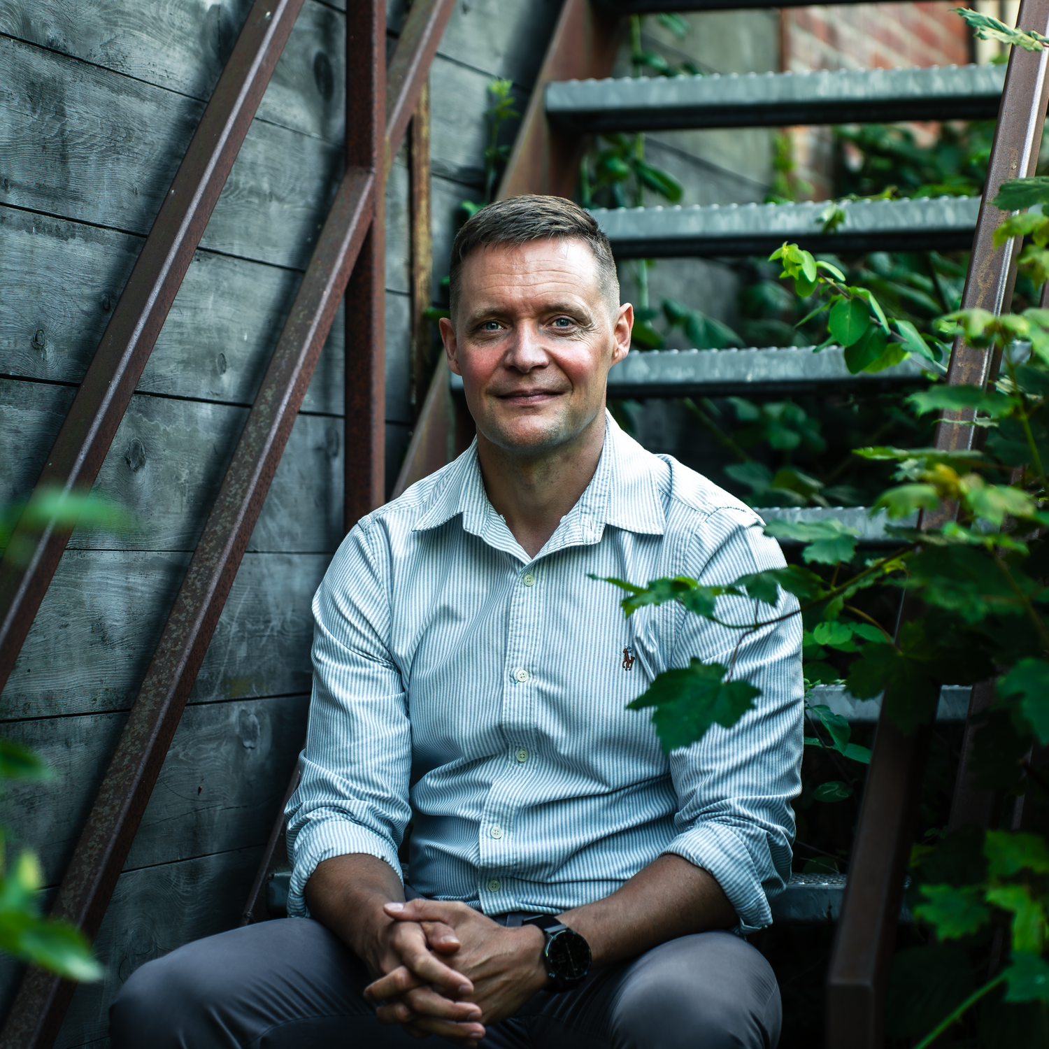 A seated person in a light blue button-up shirt smiles outdoors near green foliage and a wooden structure.