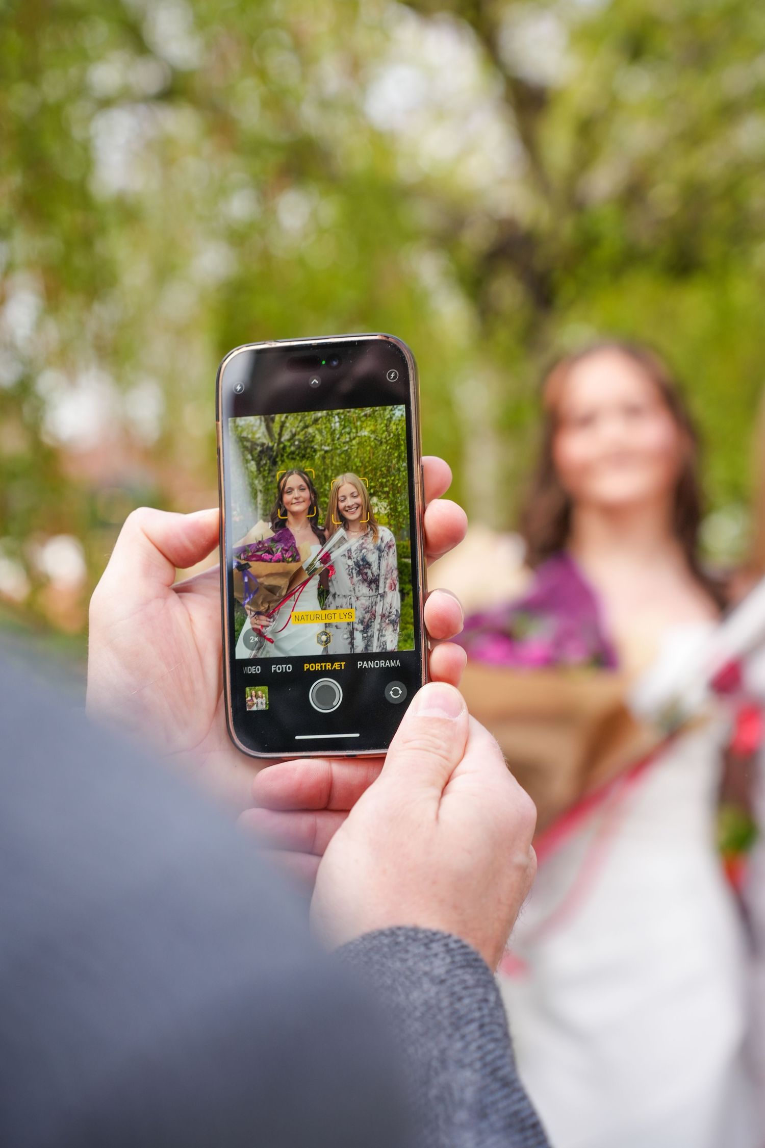Someone taking a photo with a smartphone of two people posing outdoors with green foliage in the background.
