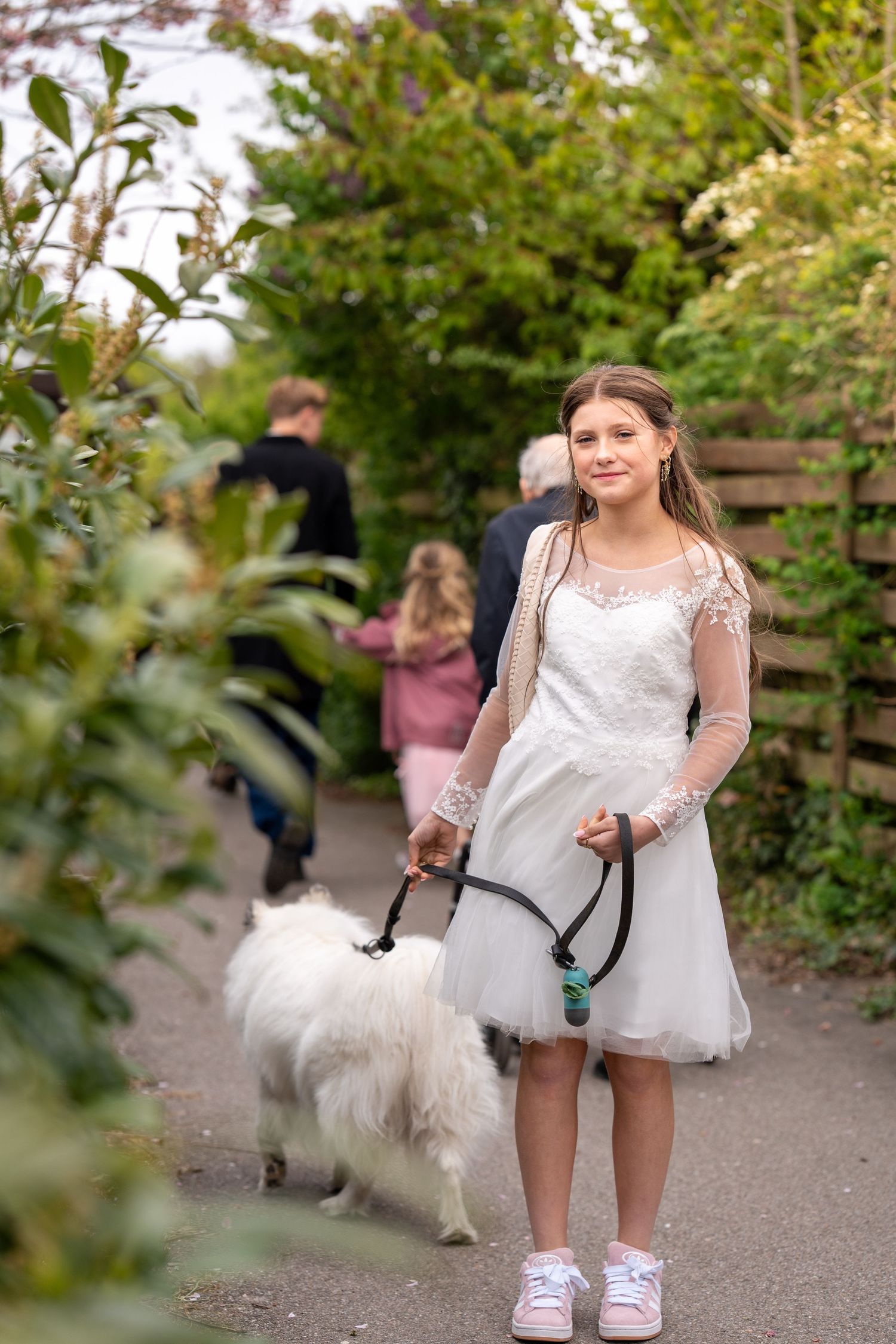 Person in a white lace dress and sneakers walking a fluffy white dog on a garden path.