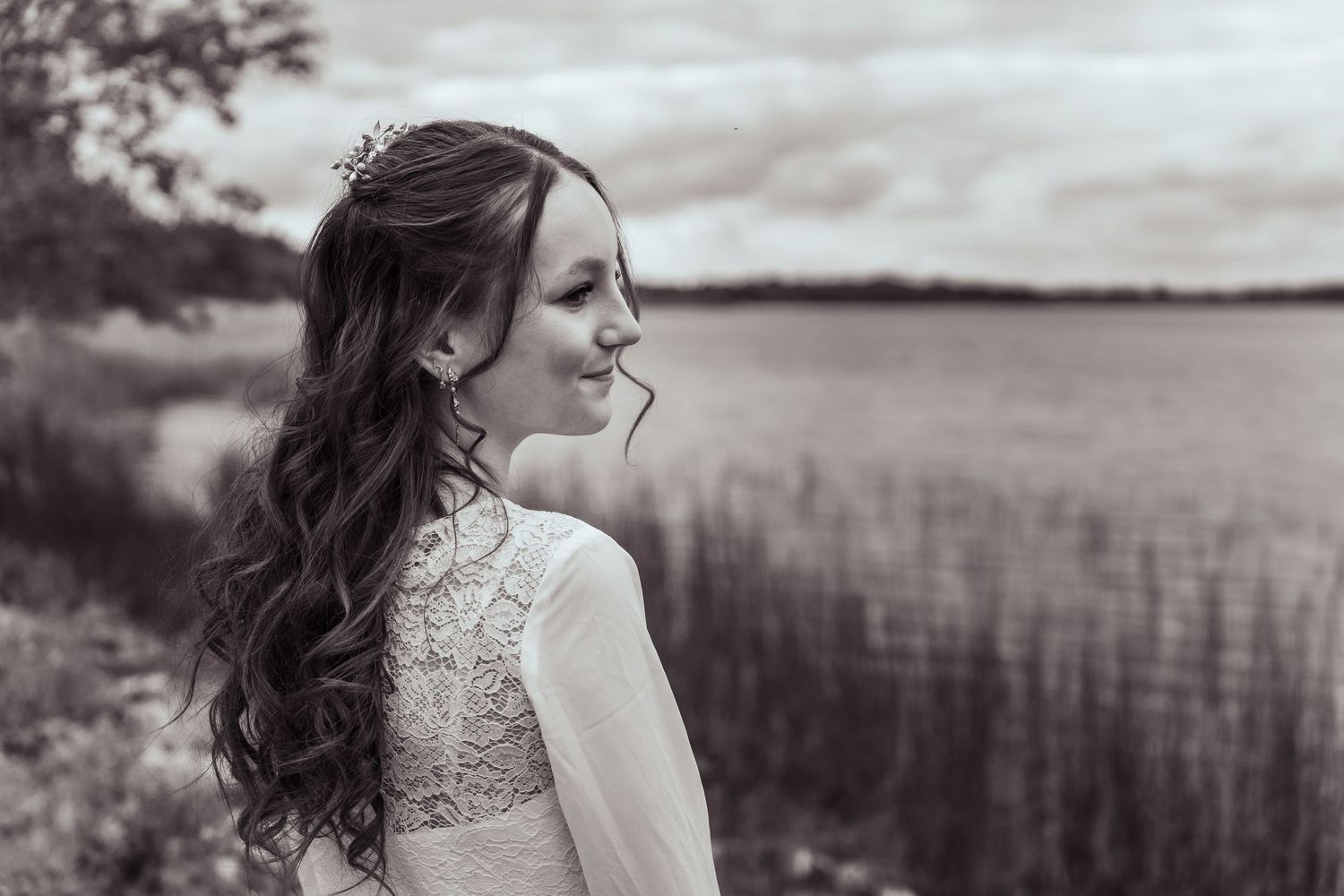 A woman in a lace wedding dress stands in profile against a serene lake landscape in black and white.