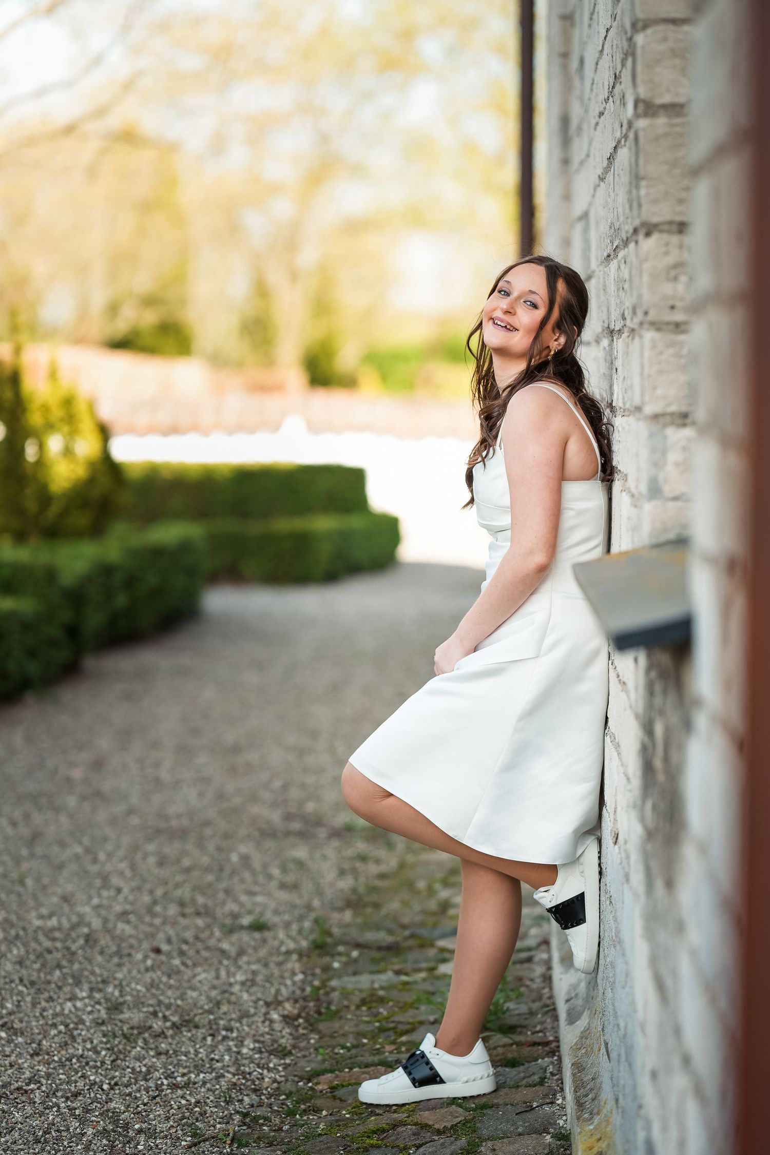 Person in a white dress and sneakers leans against a stone wall in a garden setting during golden hour.
