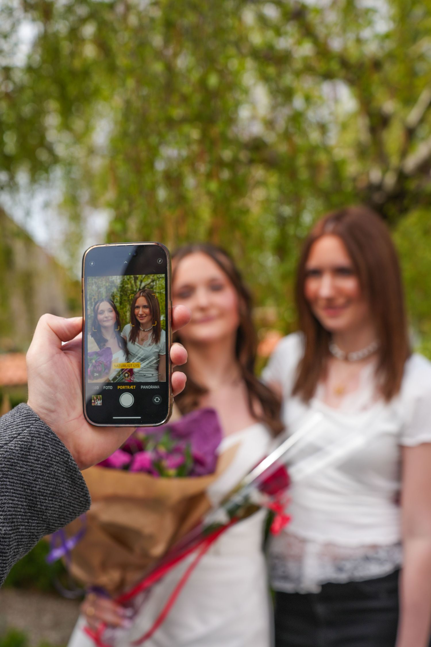 A smartphone captures a photo of two people holding bouquets in front of lush greenery outdoors.