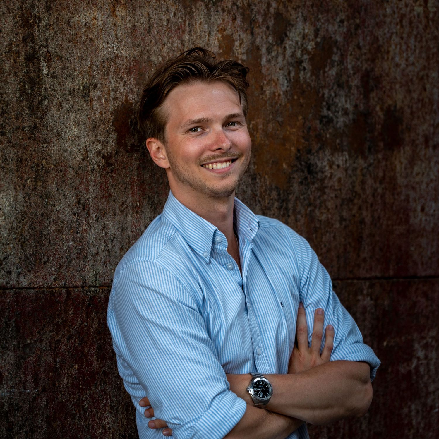 A person in a light blue button-down shirt poses expressively against a textured dark brown backdrop.