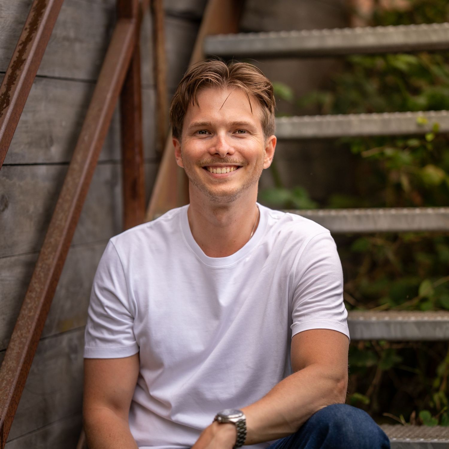 A headshot portrait in a casual outdoors setting with wooden fence backdrop, wearing a white t-shirt.
