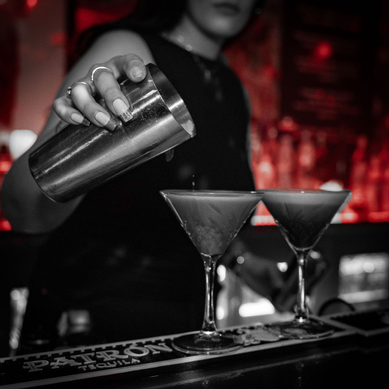 Bartender hælder cocktails i martiniglas på en mørk bar med rød stemningsbelysning.