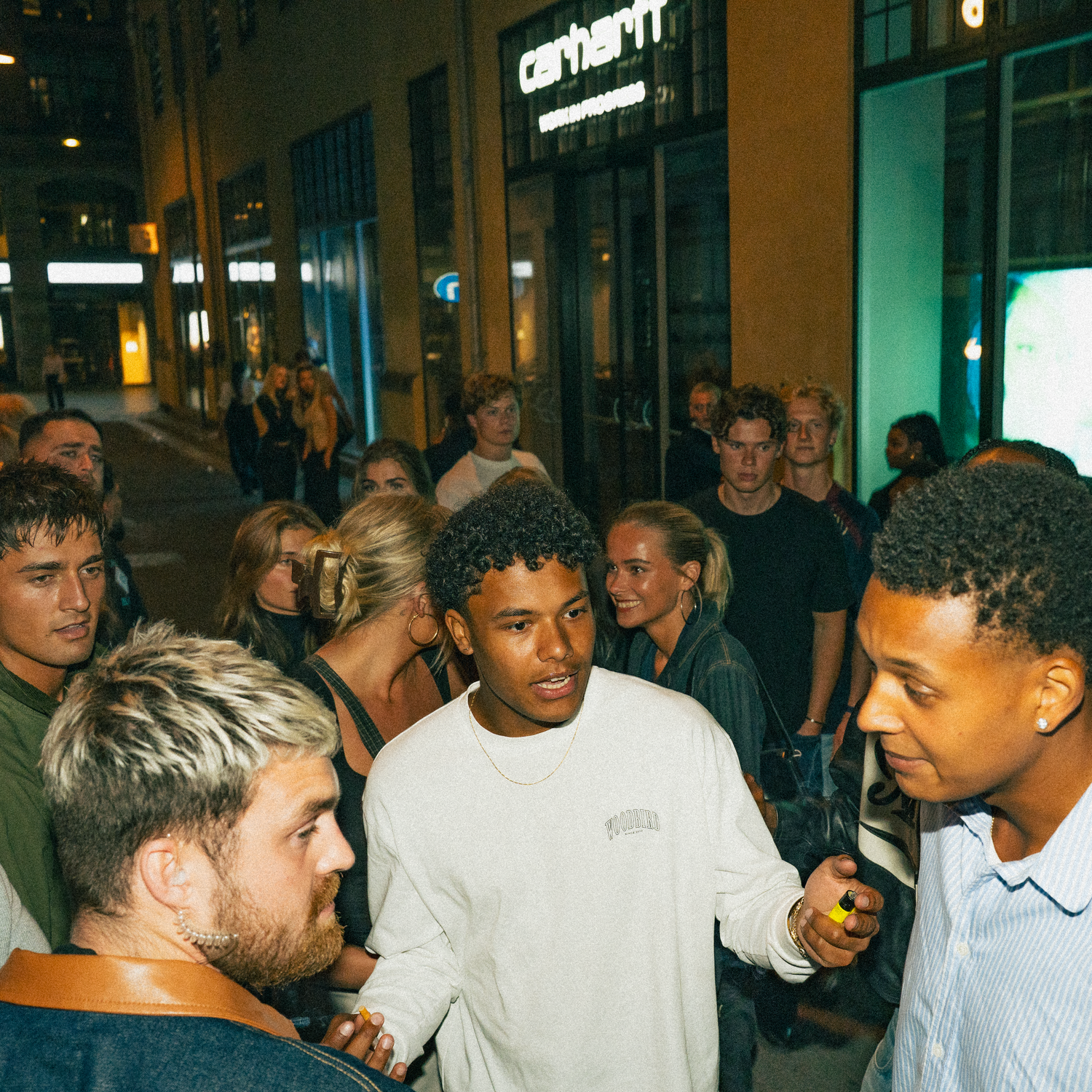 Crowd of people gathered outside at night wearing white t-shirts and casual clothing in an urban setting.